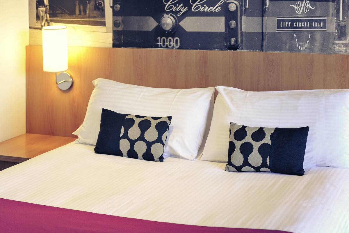 A hotel room with a bed, two pillows, a nightstand, a lamp, and a wall with a vintage train image.