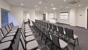 Une salle de conférence avec des rangées de chaises, un écran de projection et un pupitre orné du logo de l’hôtel Mercure North Melbourne.