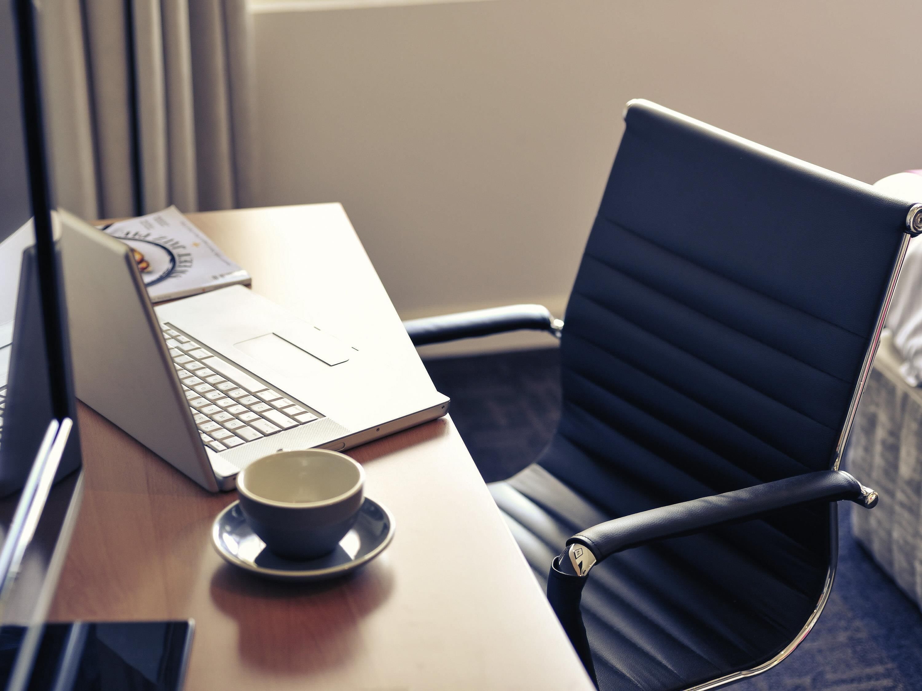 A laptop on a desk with a cup and saucer, a chair, and a window with blinds.