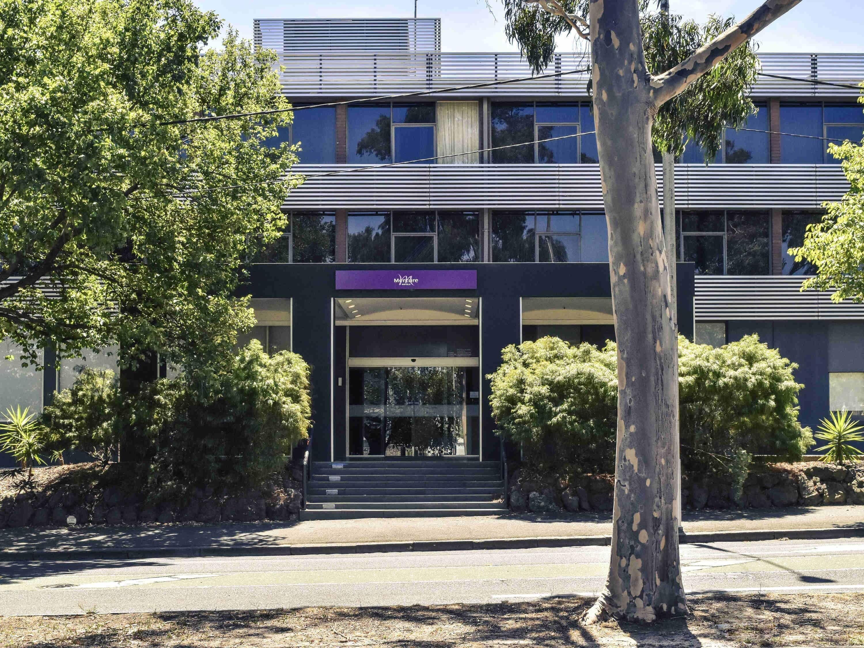 Un bâtiment moderne avec une entrée en verre, entouré d’arbres et sous un ciel dégagé.