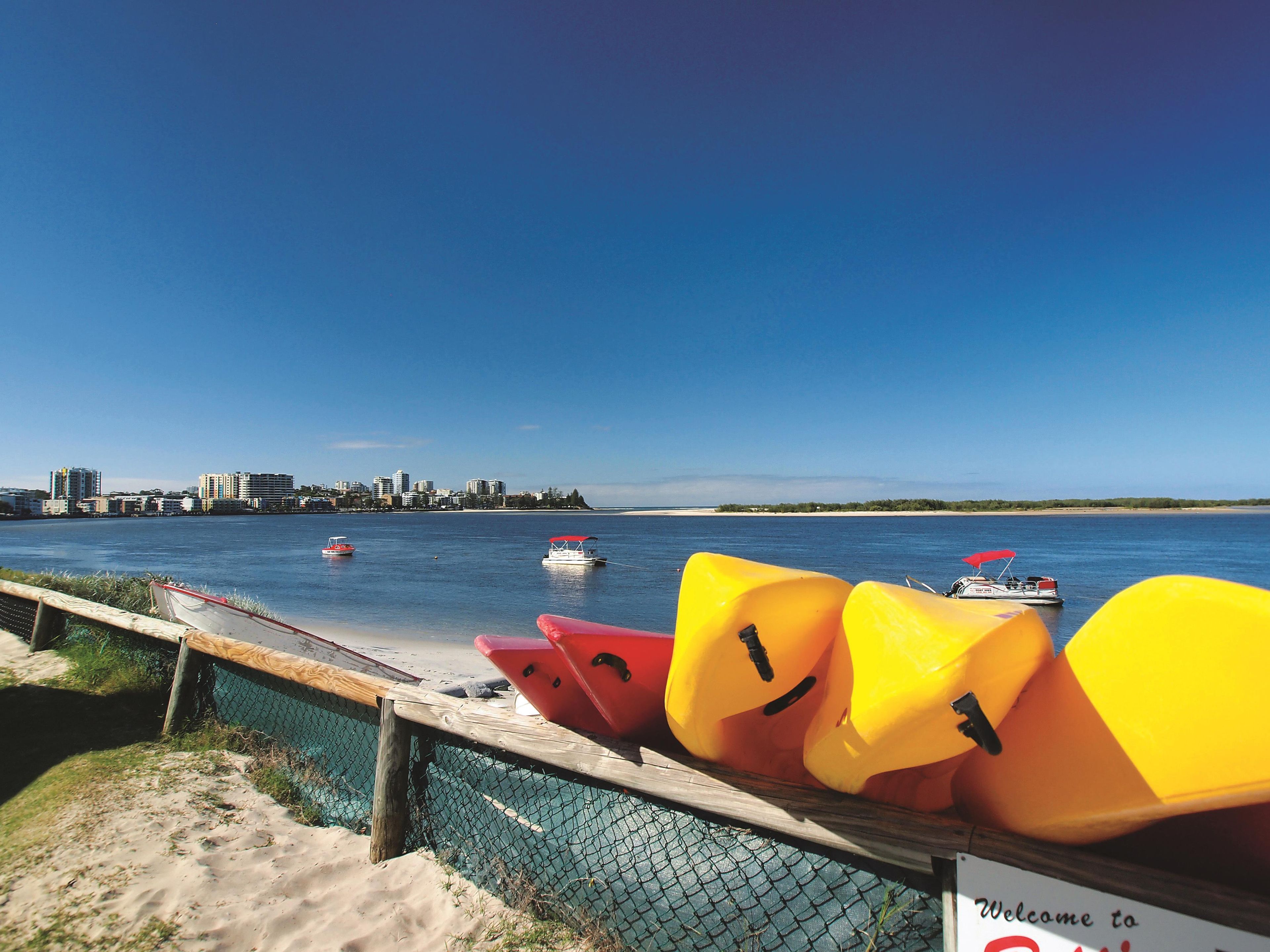 A beachside view with kayaks lined up, a signboard, and a city skyline in the background.