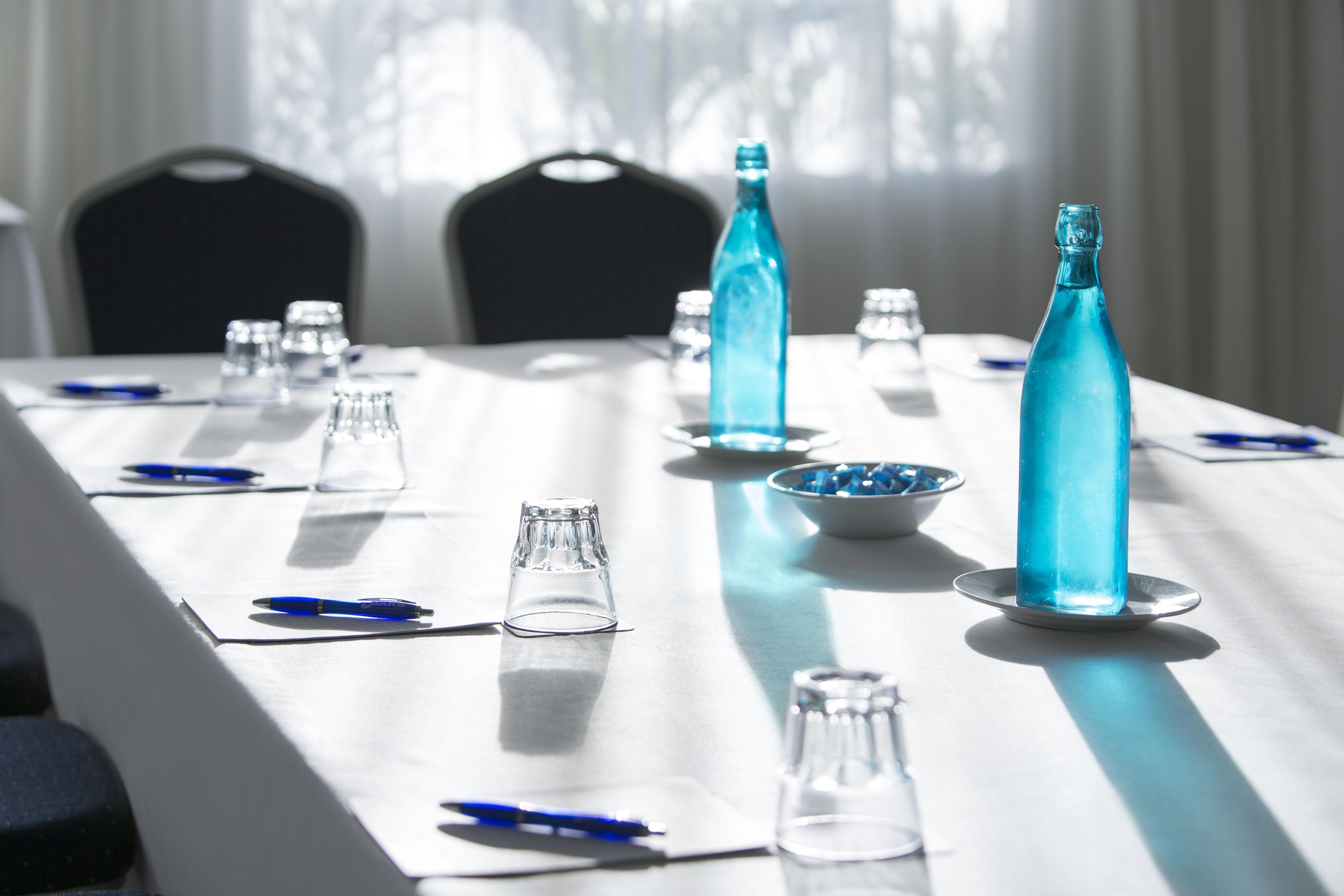 Een vergadertafel met helder glazen servies, blauwe flessen en pennen, in een ruimte met stoelen en een raam met gordijnen.