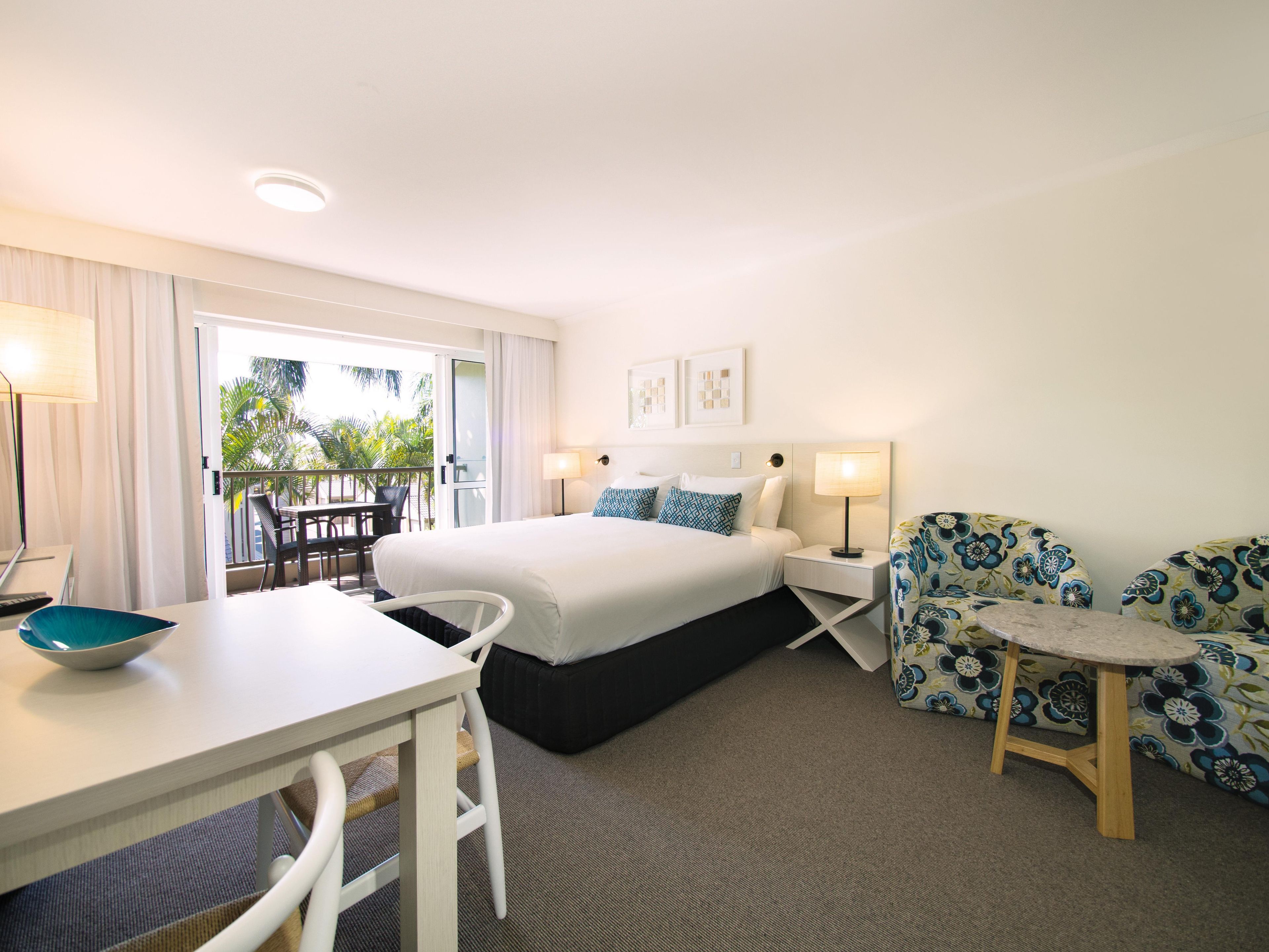Habitación de hotel con cama, escritorio, silla, una mesita y balcón con vista a palmeras.