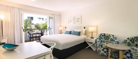 Habitación de hotel con cama, escritorio, silla, una mesita y balcón con vista a palmeras.