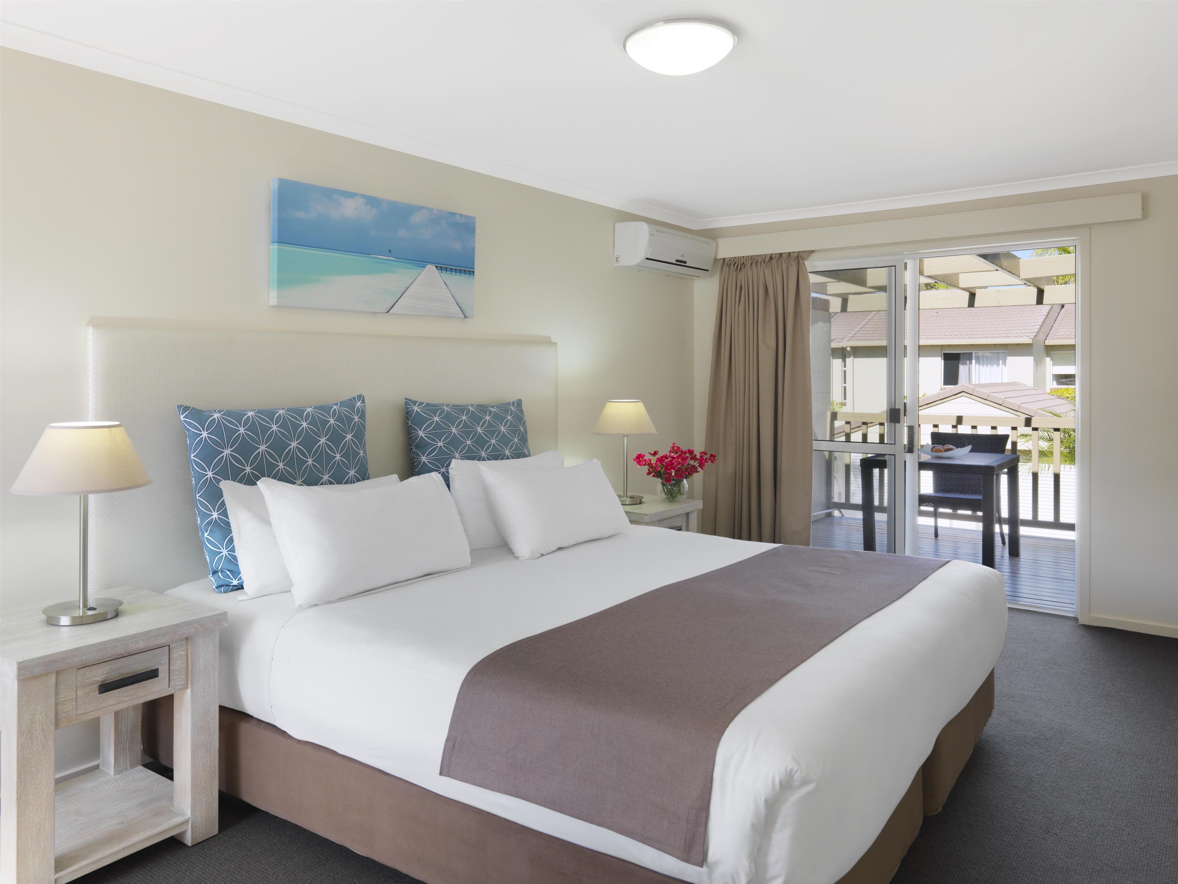 A hotel room with a bed, bedside tables, a lamp, a painting, a window with curtains, and a view of an outdoor area.