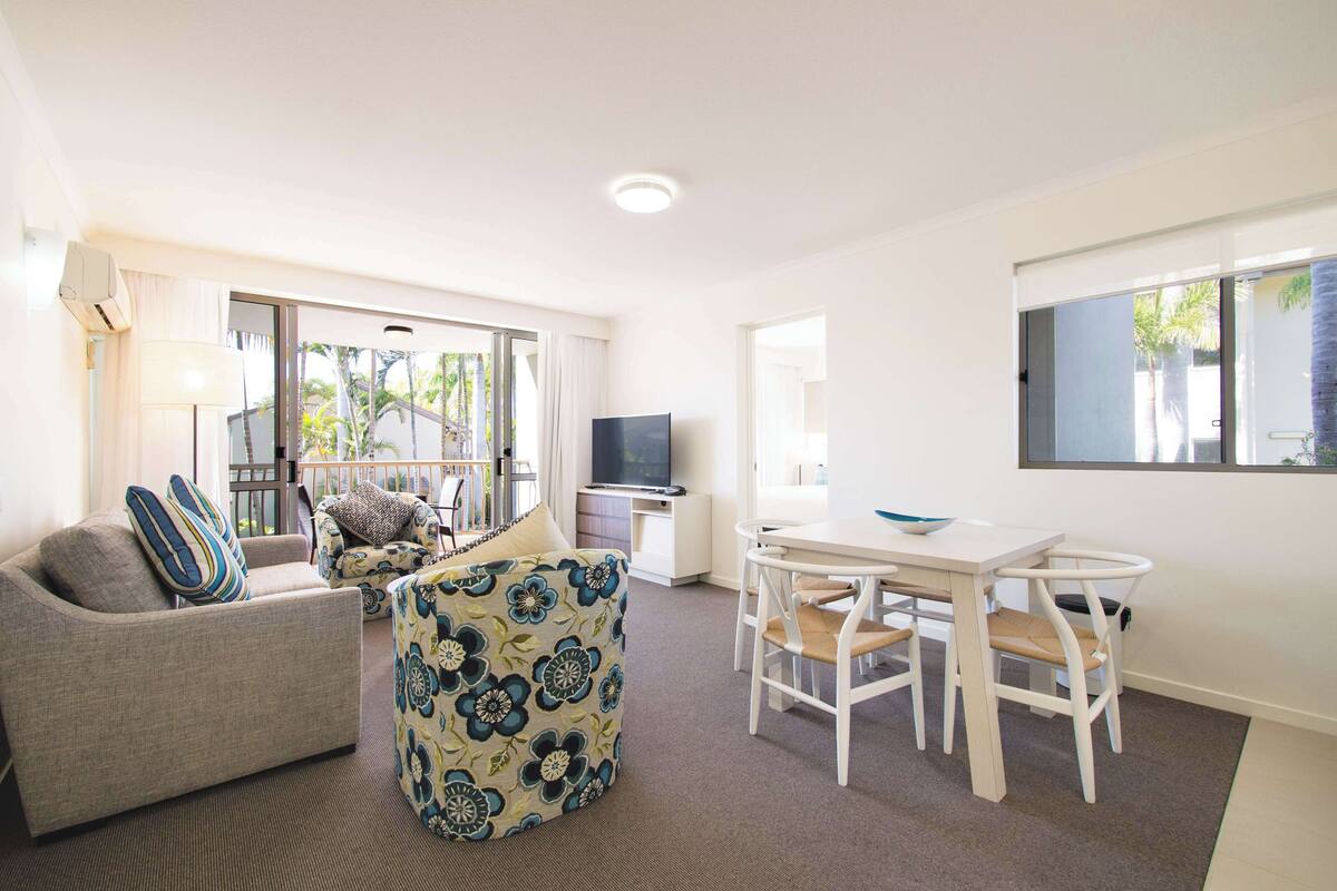 A living room with a sofa, a patterned ottoman, a dining table with chairs, a flat-screen TV, and large windows with a view of palm trees.