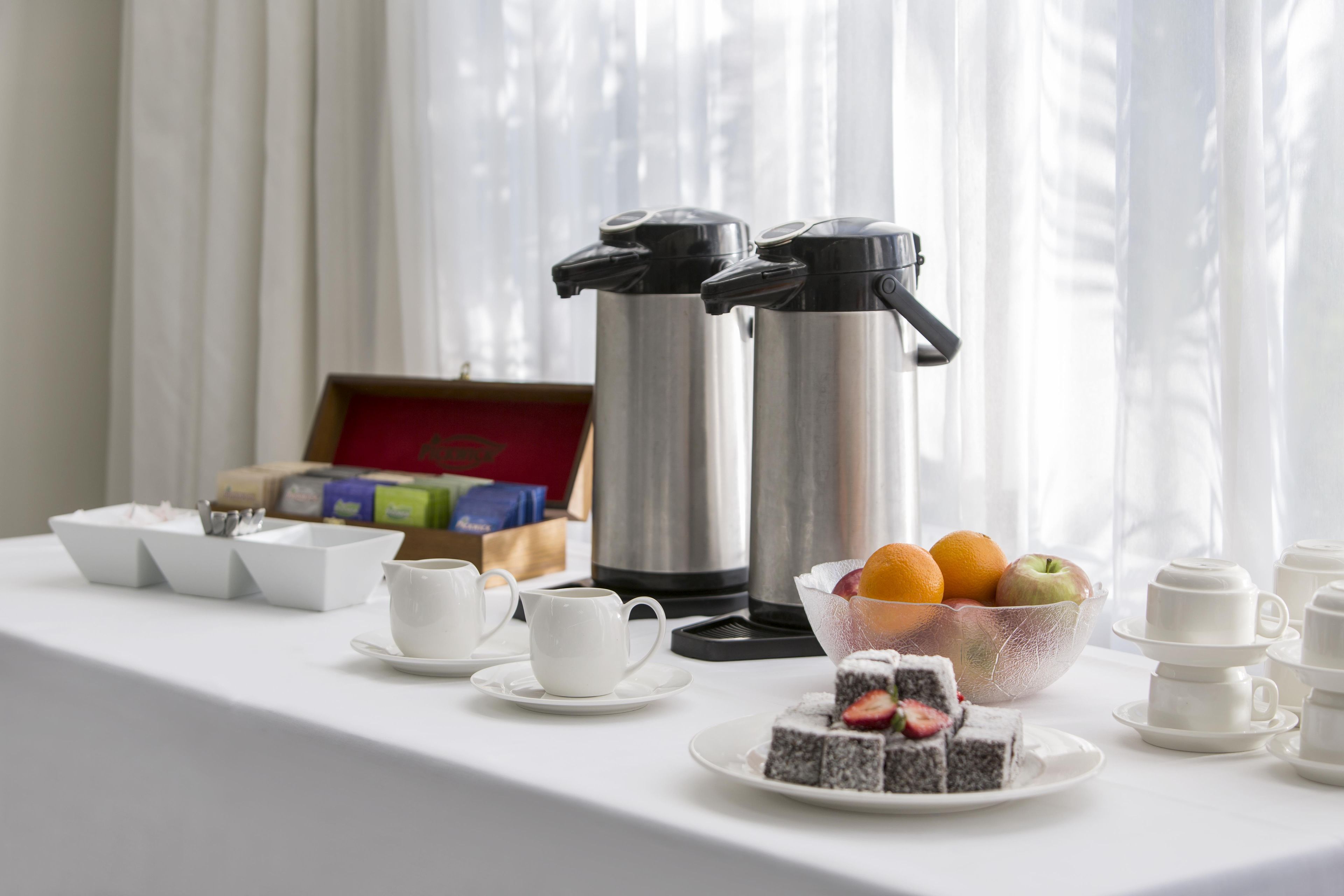 A breakfast setup with coffee makers, cups, a bowl of fruit, and a plate of cake.