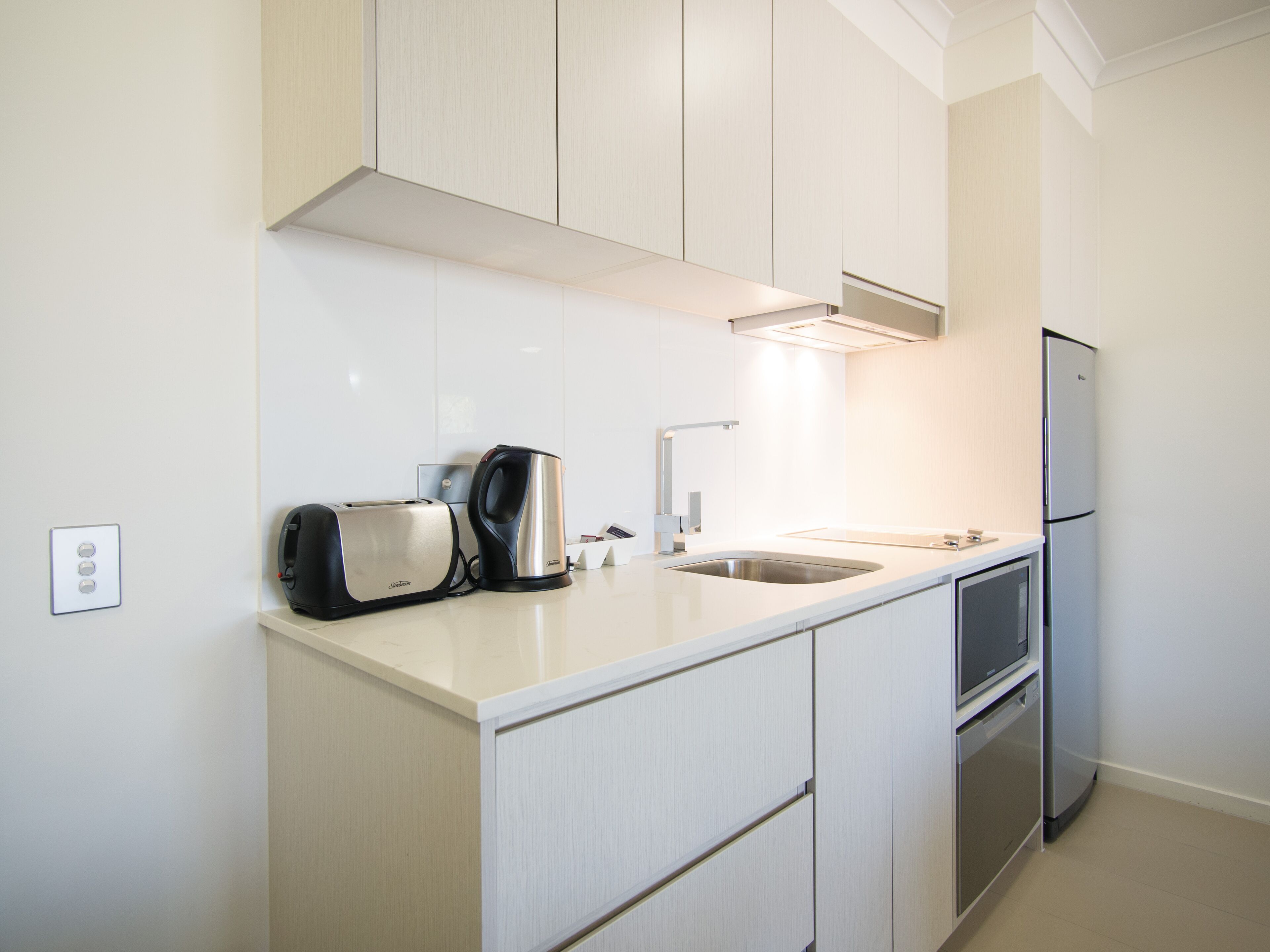 A modern kitchen with white cabinetry, a stainless steel toaster, a kettle, a sink, and a refrigerator.