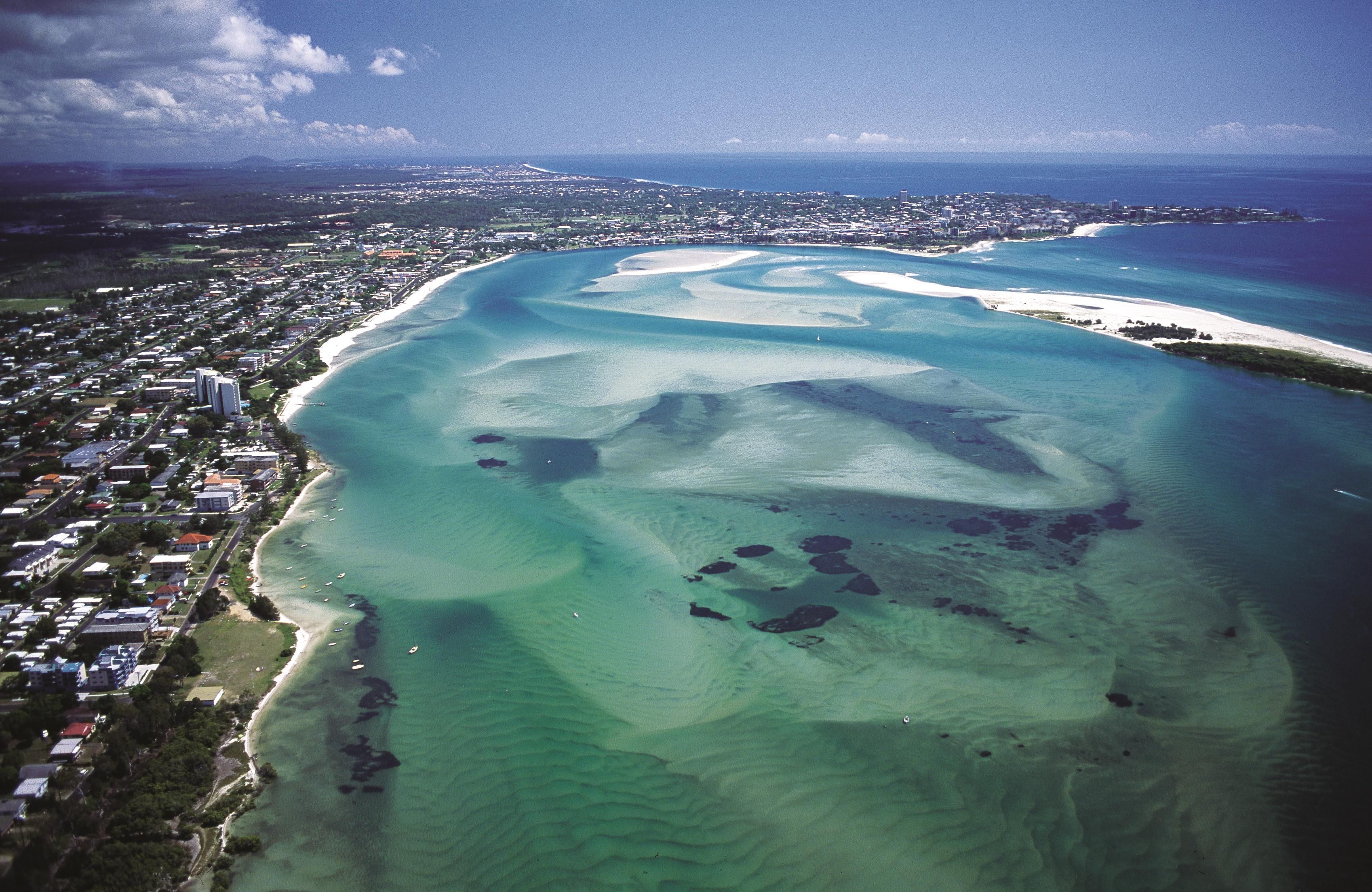 An aerial view of a coastal town with a beach, clear waters, and a distant city skyline.
