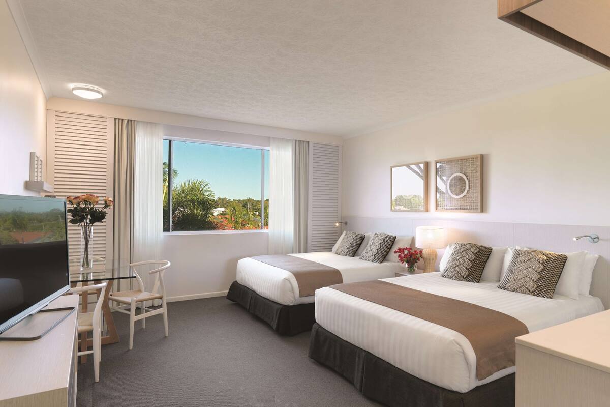 A hotel room with two beds, a desk with a TV, a chair, and a window with a view of greenery.