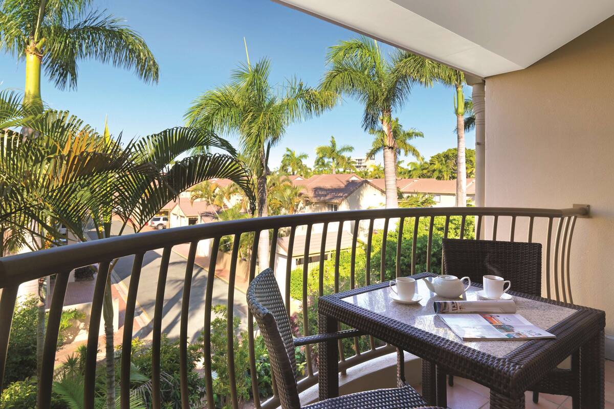 A balcony with wicker furniture, a table with cups and a newspaper, and a view of palm trees and houses.