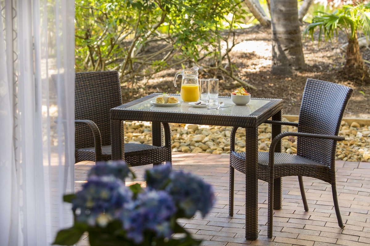 A wicker table with two chairs, a pitcher of orange juice, a glass, and a bowl of fruit on a patio with a brick floor and greenery in the background.