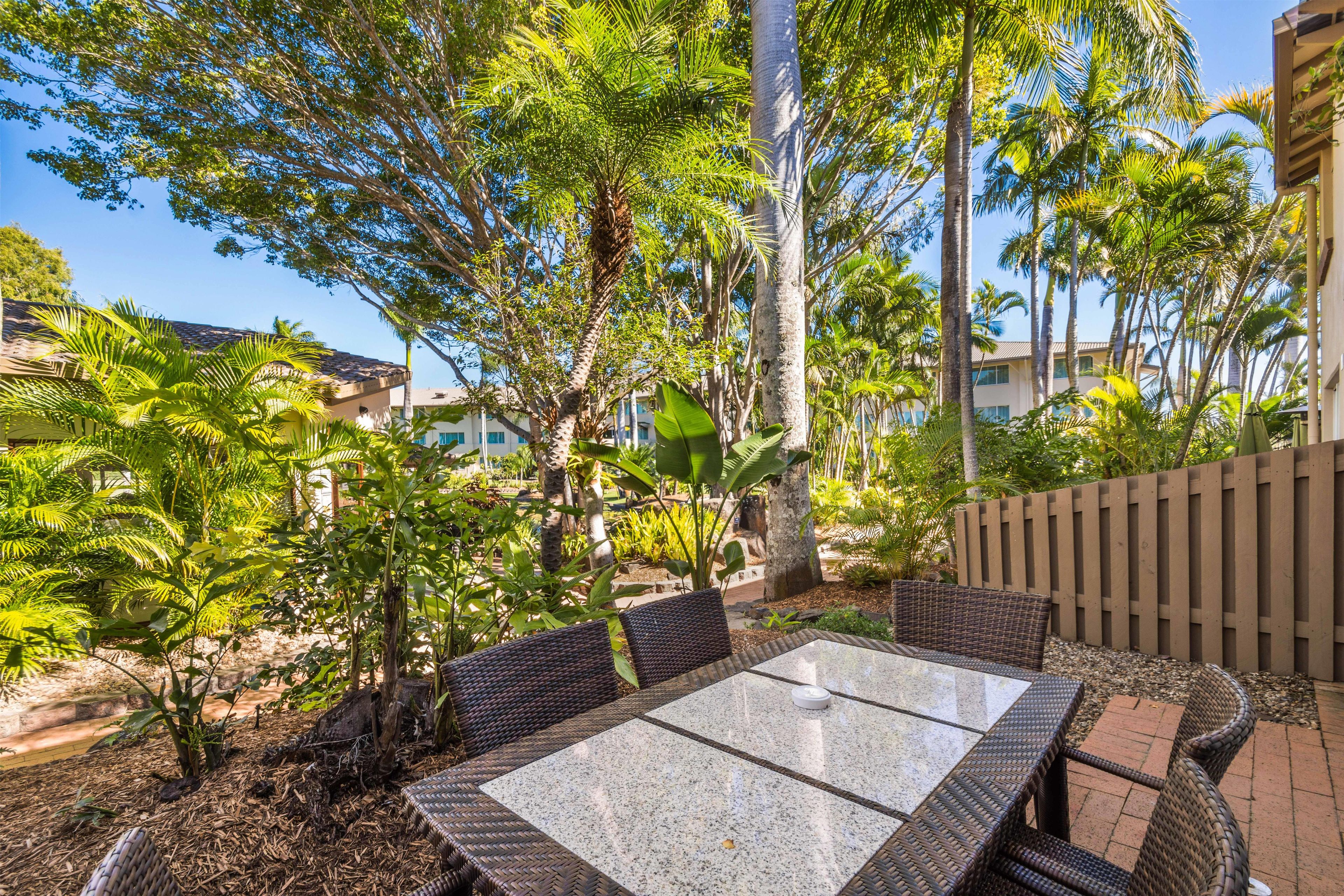 An outdoor patio area with wicker furniture and a tiled table, surrounded by lush greenery and tall trees.