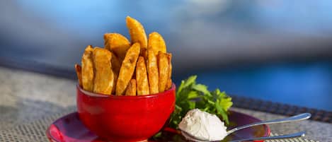 Eine Schüssel goldbraune Pommes auf einem roten Teller mit einem Löffel, daneben eine Portion weißer Dip, auf einem Tisch mit unscharfem Hintergrund.