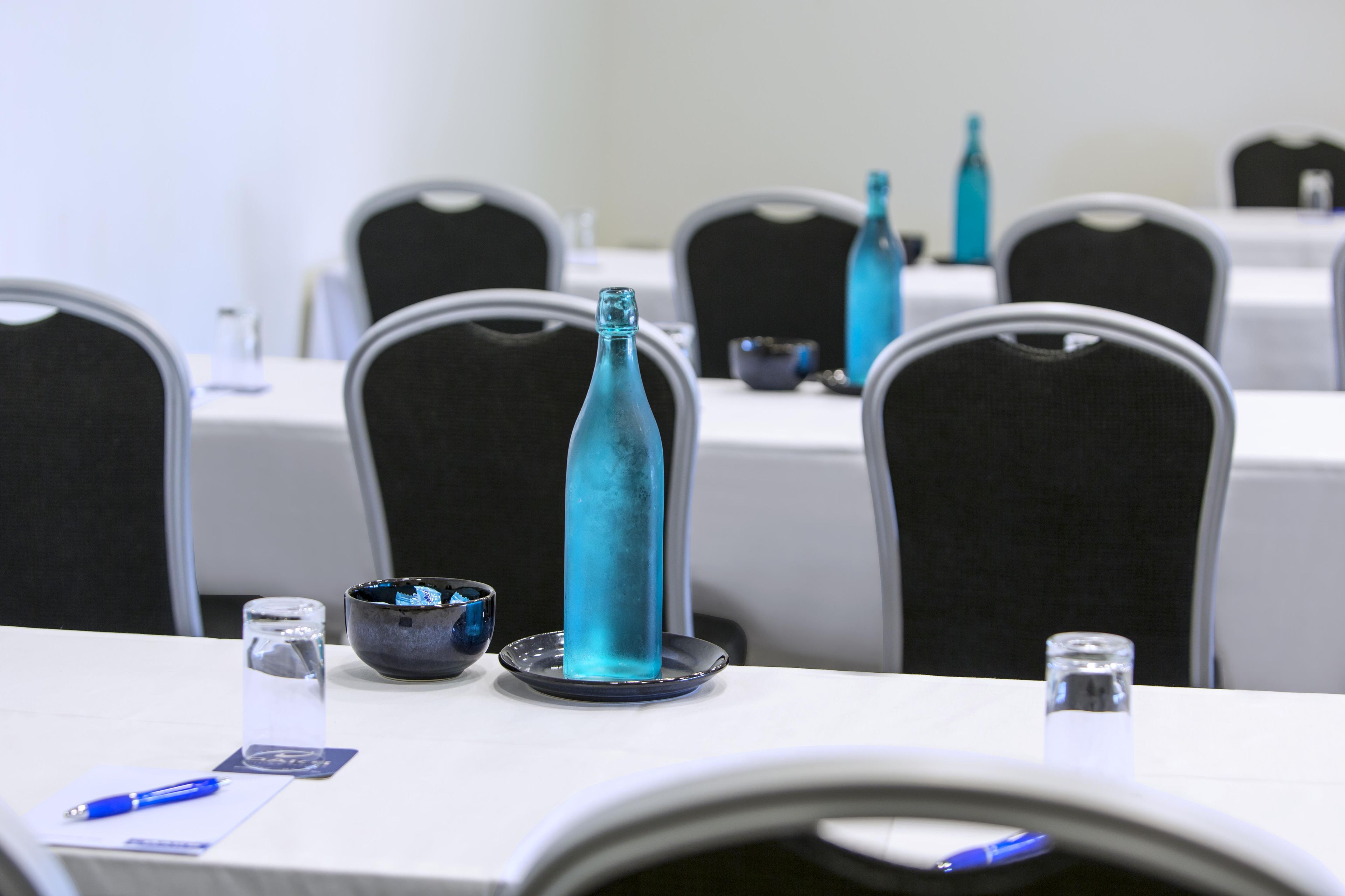Un salón de conferencias con mesas dispuestas para reuniones, equipado con tarjetas de identificación, bolígrafos y un vaso de agua.