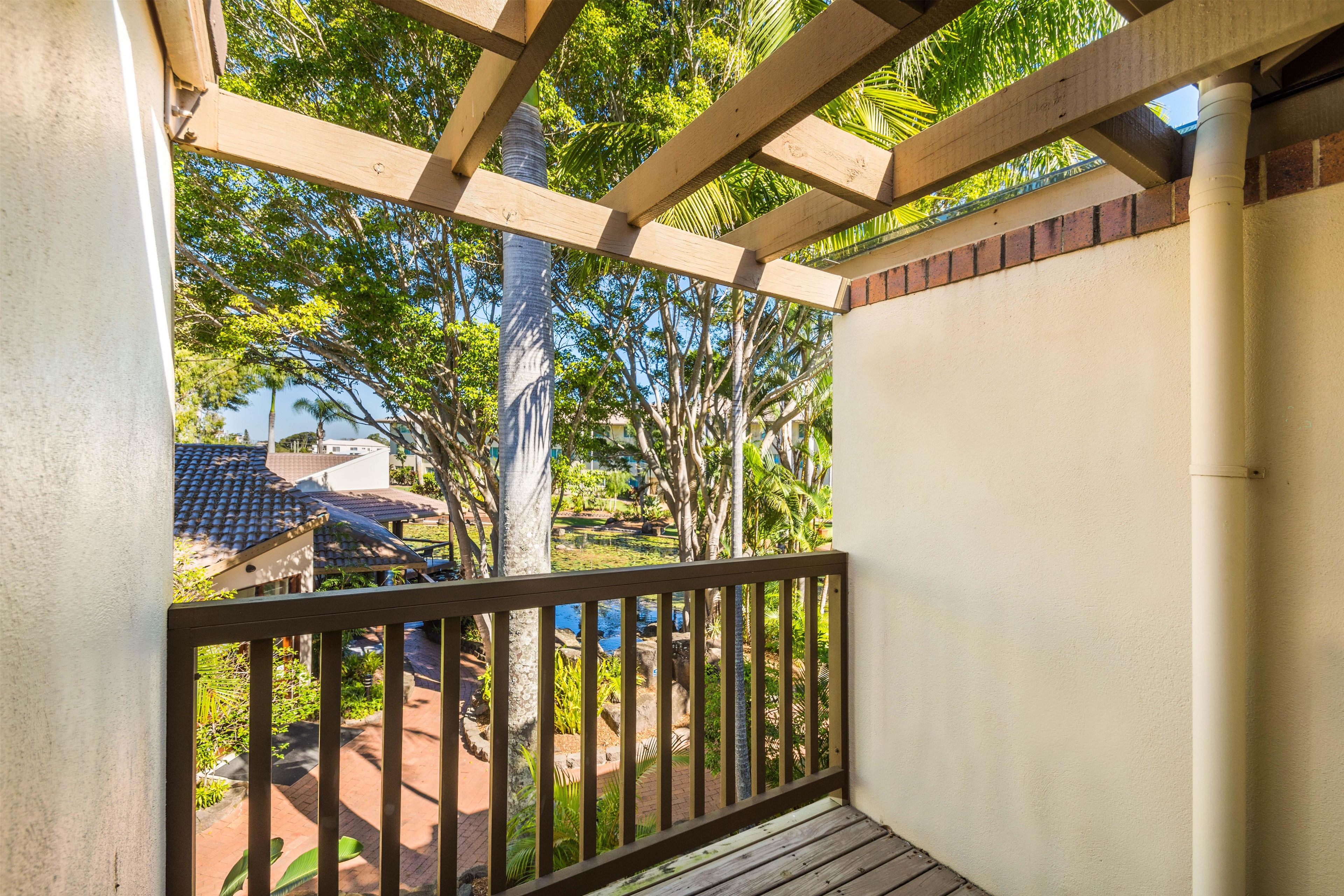Un espacio exterior cubierto con barandilla de madera, pared de hormigón y vistas a un jardín con árboles y un estanque.