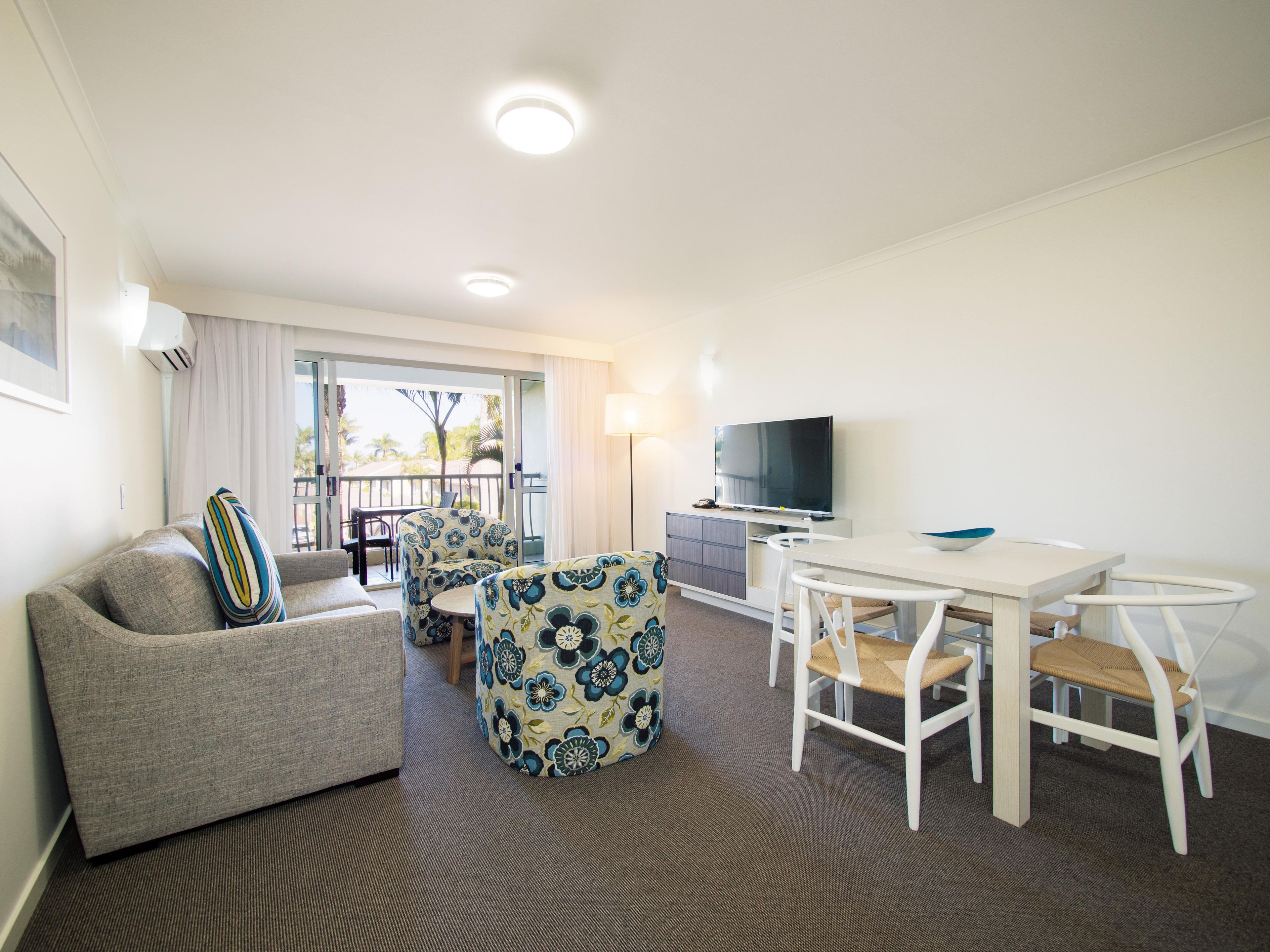A hotel room with a sofa, a dining table with chairs, a TV, and a balcony with a view.