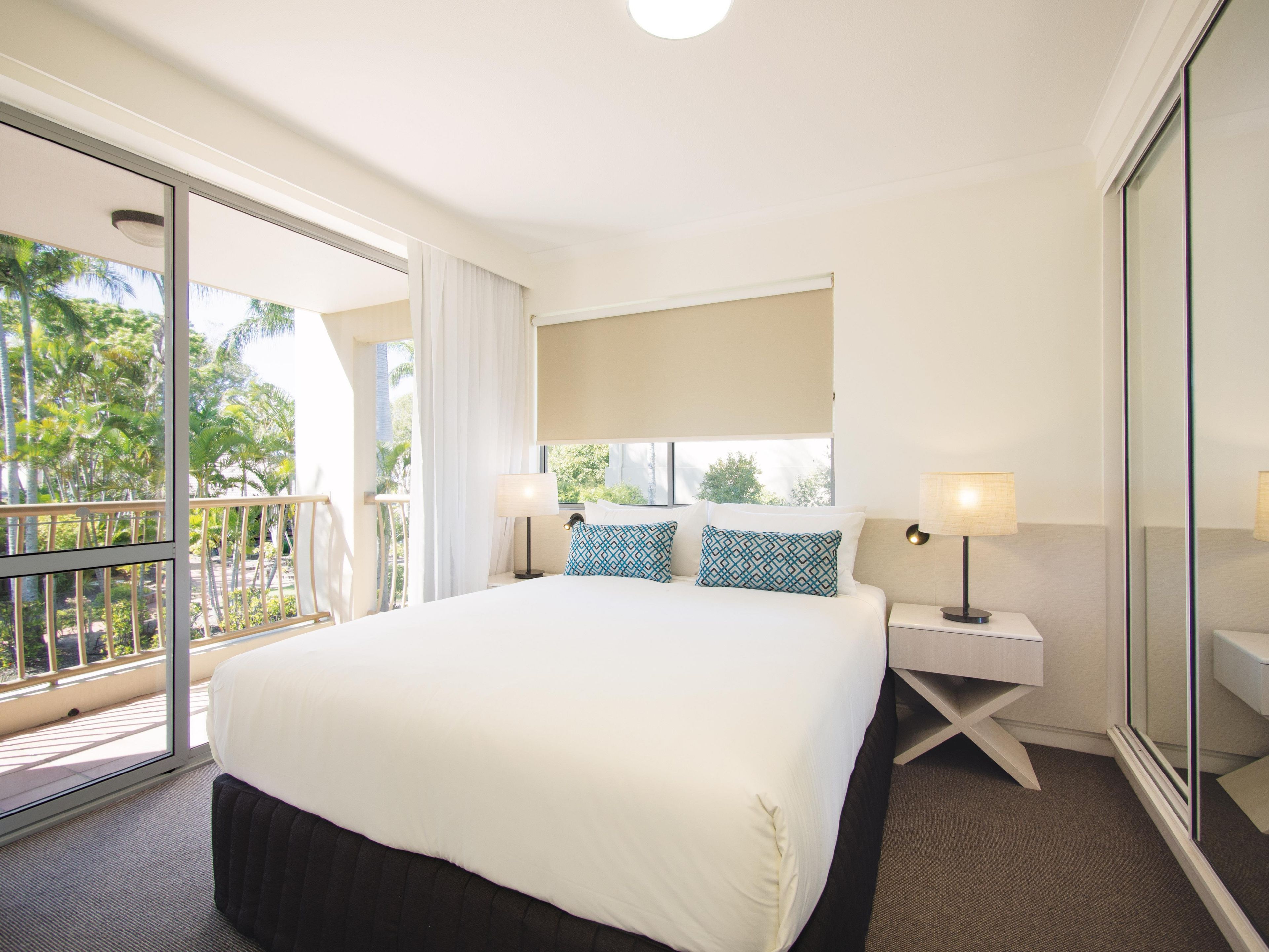 Habitación de hotel con una cama grande, dos mesitas de noche con lámparas, un ventanal con vistas a palmeras y un balcón.