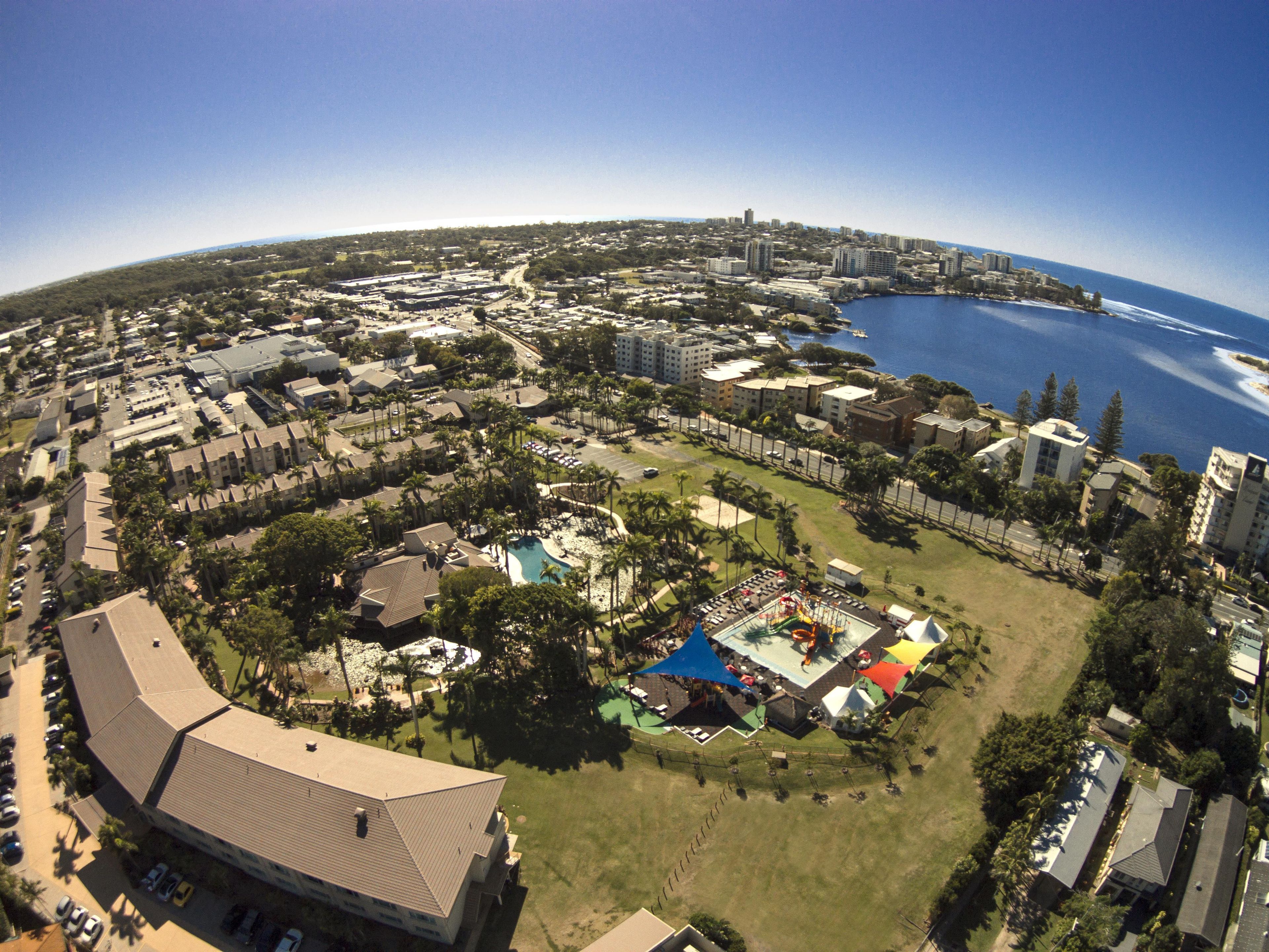 An aerial view of a coastal city with a mix of residential and commercial buildings, a marina, and a large public park with a playground.