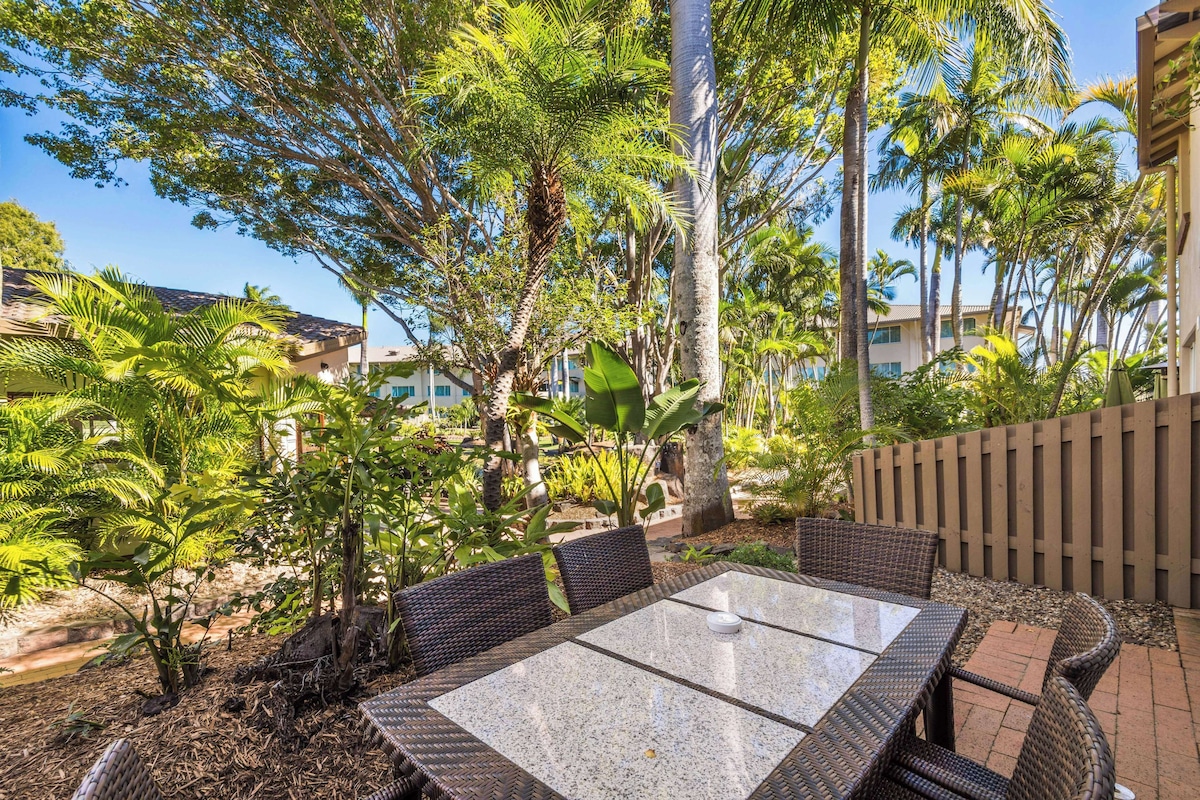 An outdoor patio area with a glass-top table surrounded by wicker chairs, set against a backdrop of lush greenery and tall palm trees.