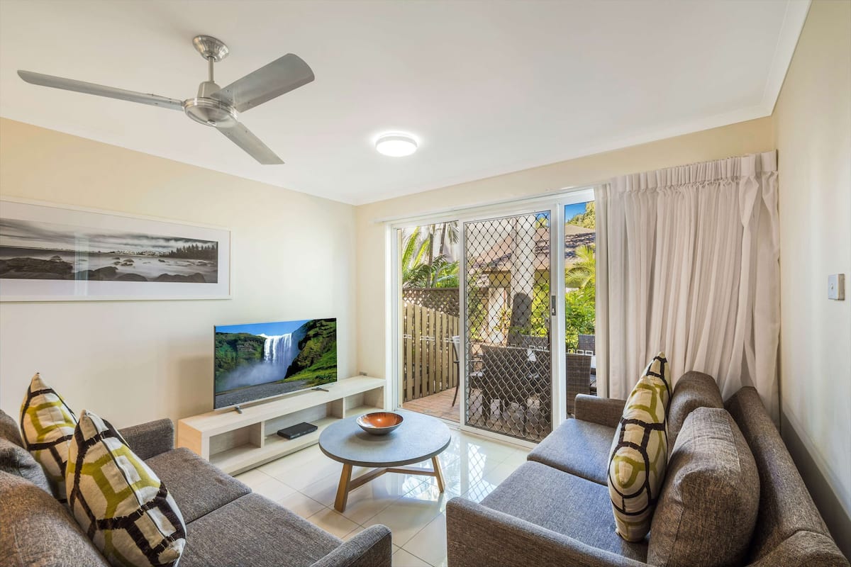 A living room with a flat-screen TV, grey sofas, a coffee table, and a ceiling fan.