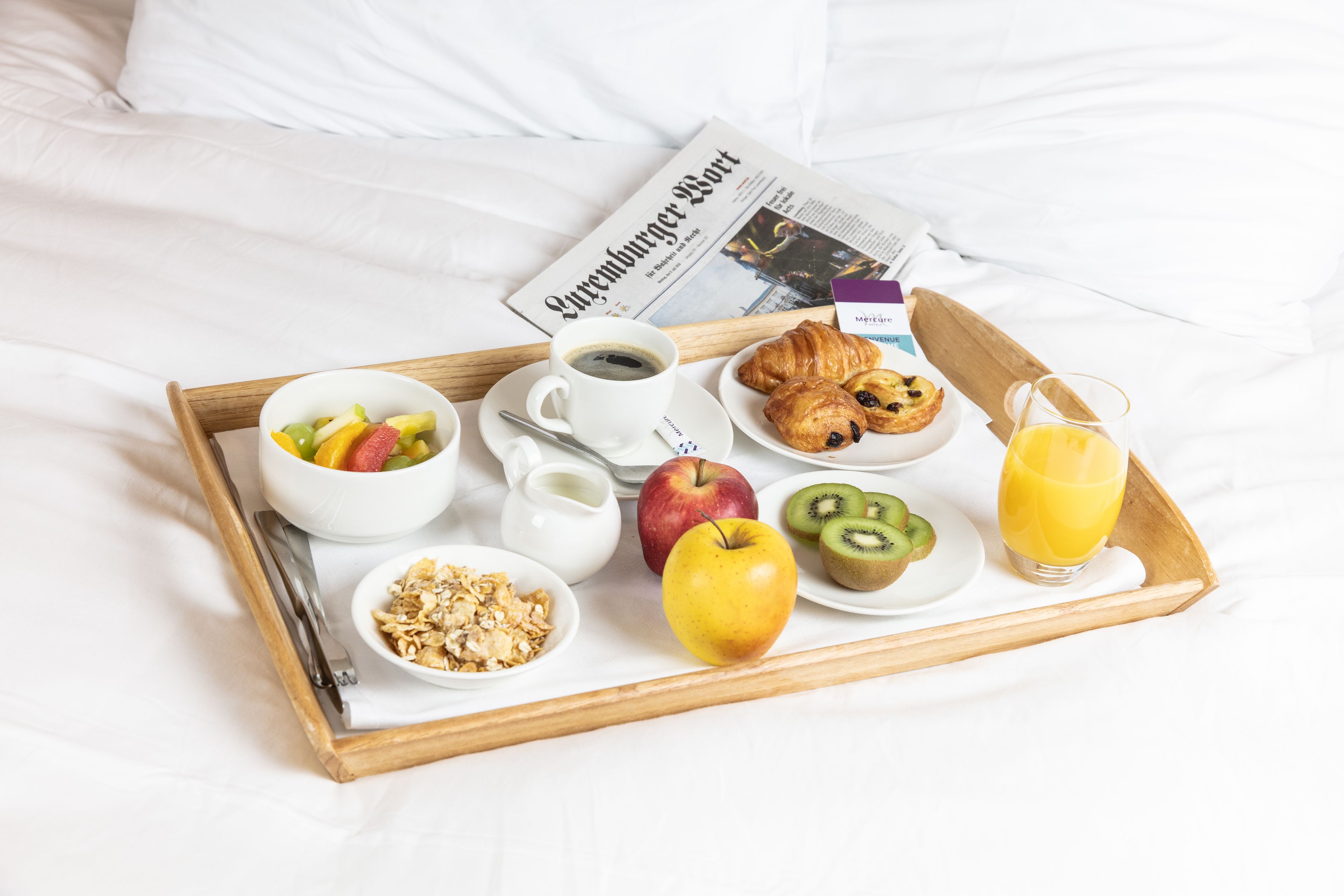 Una bandeja de desayuno con frutas, cruasanes, jugo y un periódico sobre la cama.