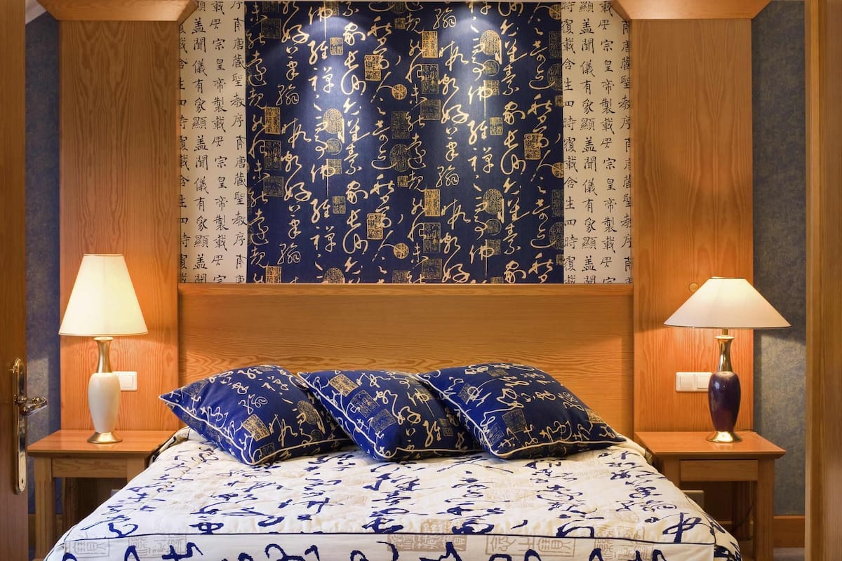 A bedroom with a bed, two bedside lamps, a wooden headboard, and a wall with calligraphy.