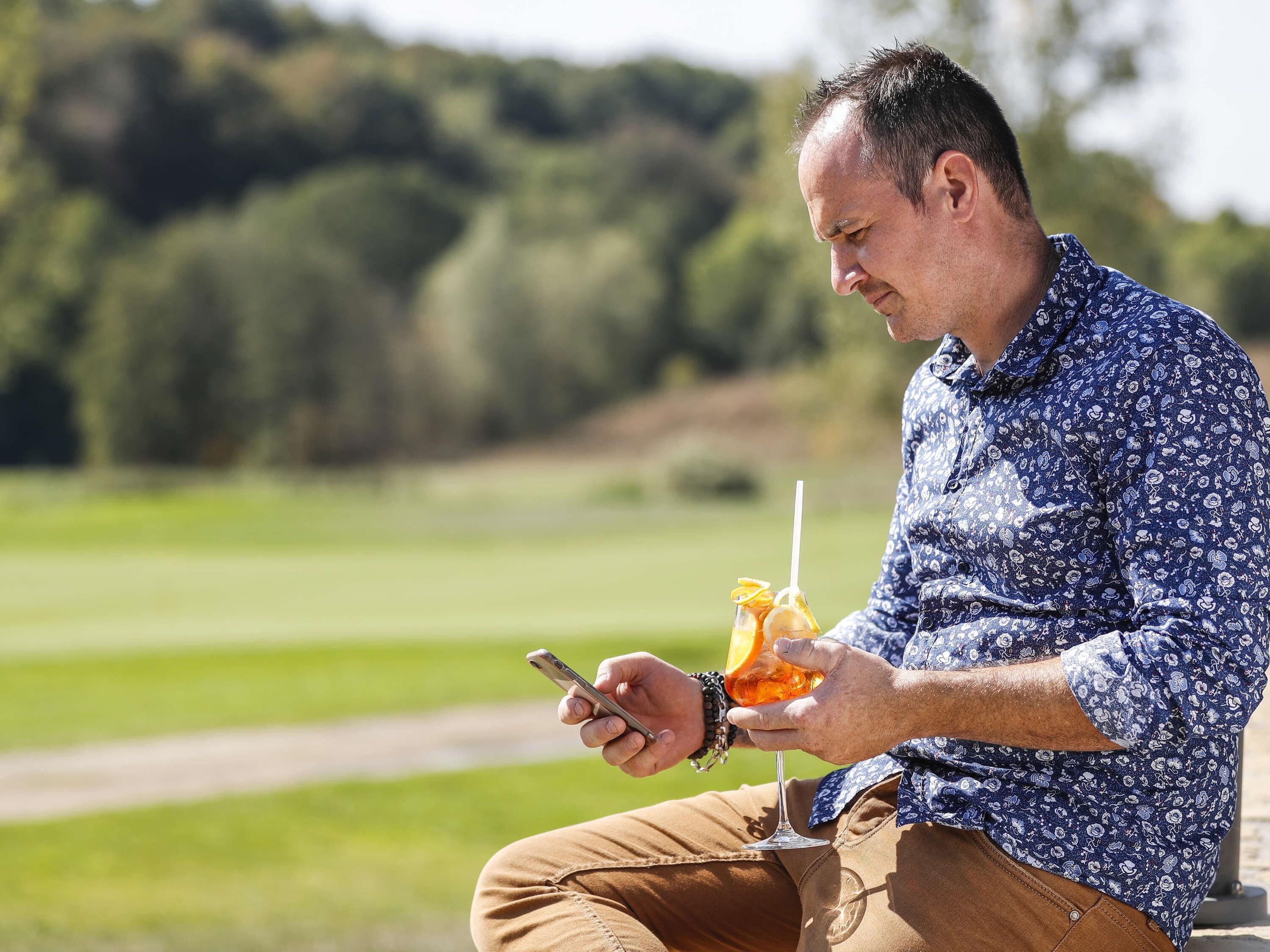 Eine Person sitzt draußen, hält ein Smartphone und ein Getränk mit Orangenscheibe.