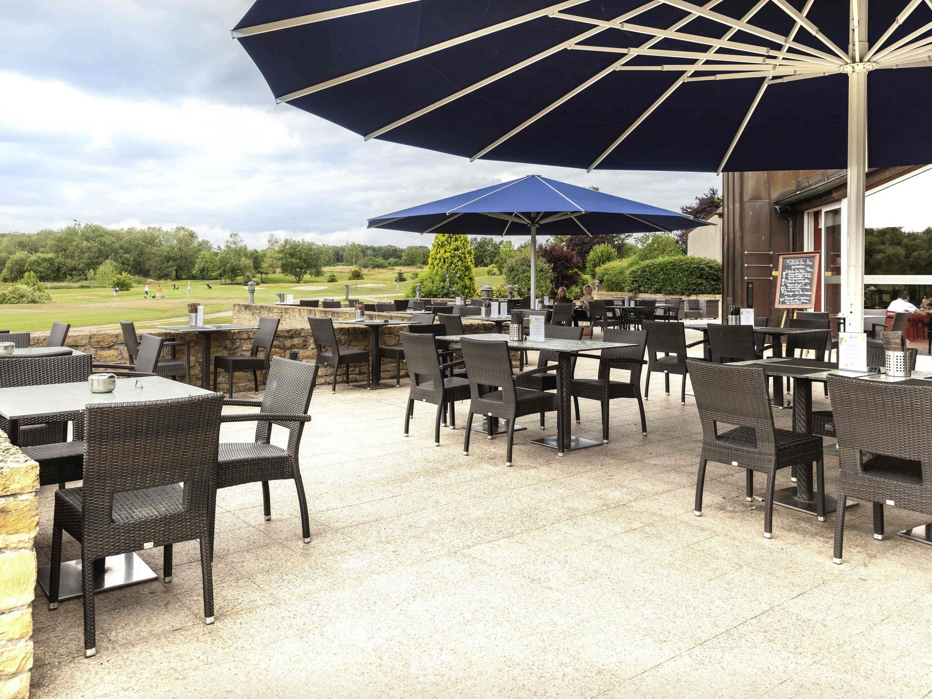 Een terras buiten met tafels en stoelen, een grote parasol en uitzicht op een golfbaan.
