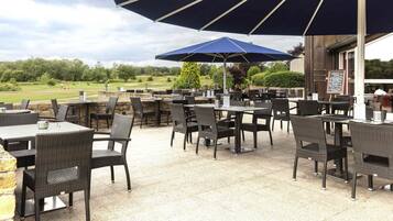 Een terras buiten met tafels en stoelen, een grote parasol en uitzicht op een golfbaan.