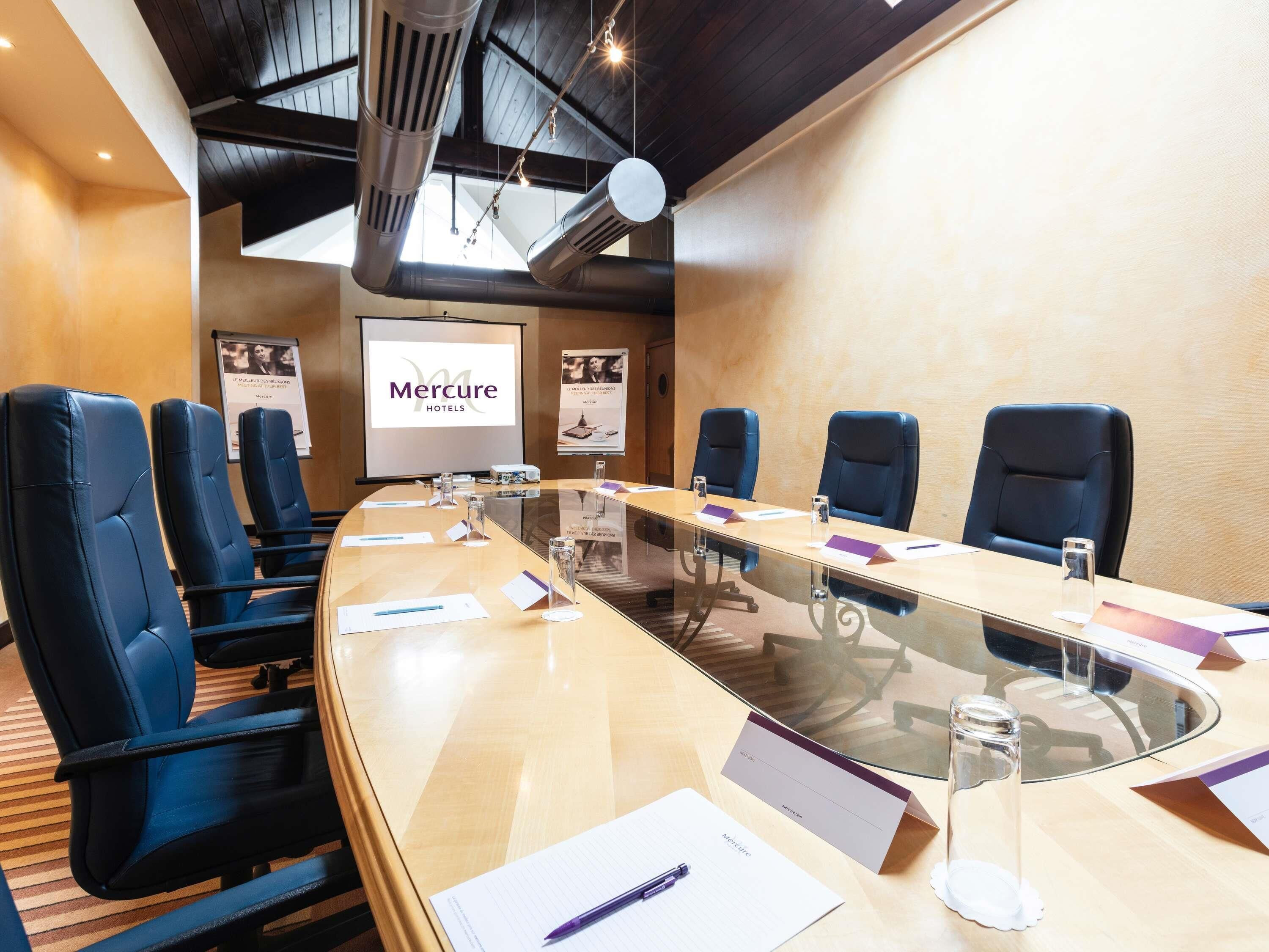 A conference room with a long table, blue chairs, a projector screen, and a 'Mercure Hotels' sign.
