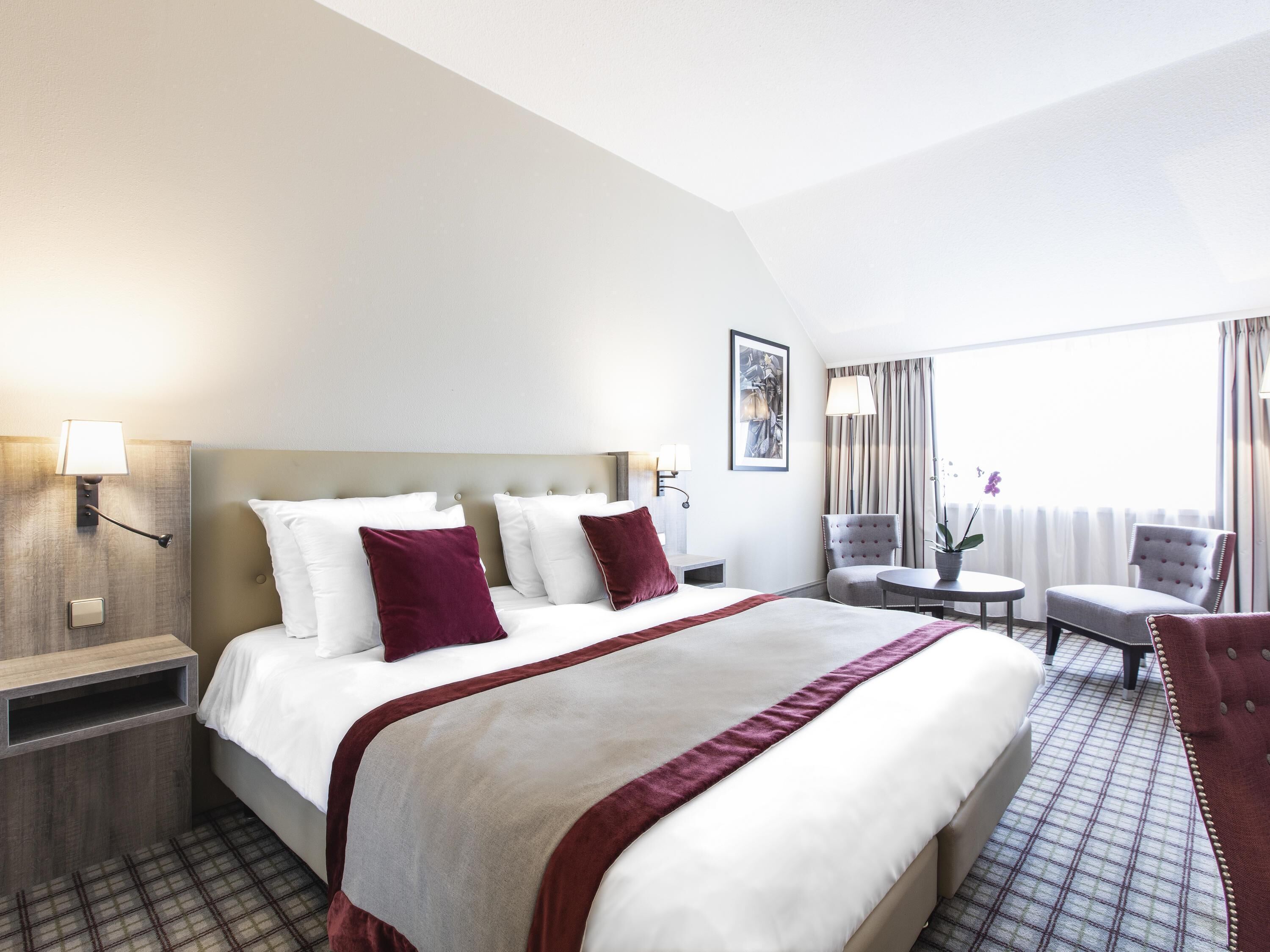 A hotel room with a large bed, two bedside tables, a chair, a small table, and a window with curtains.