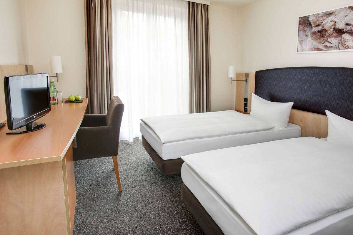 Een hotelkamer met twee bedden, een bureau met een televisie, een stoel en een raam met gordijnen.