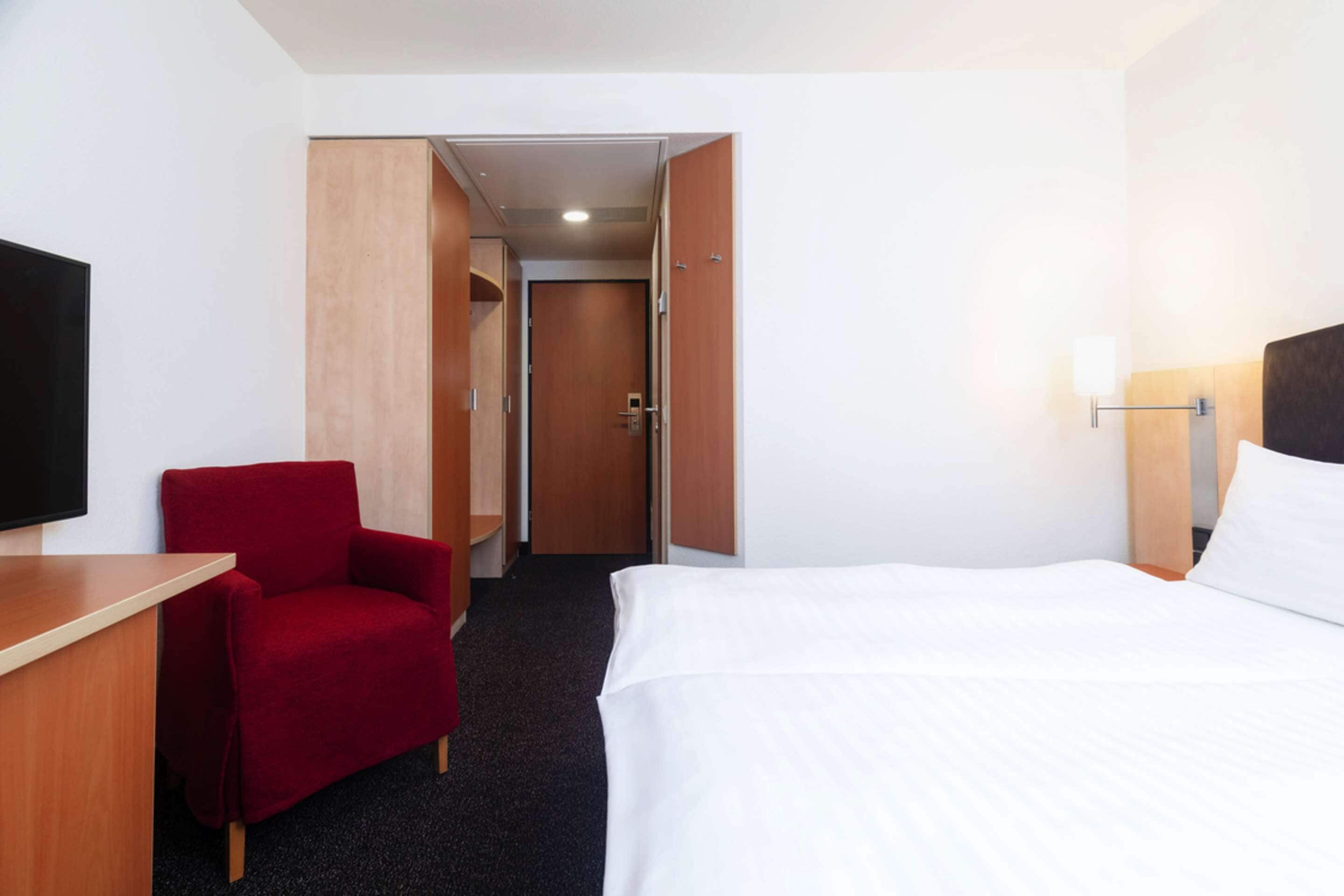 A hotel room with a bed, a red armchair, a desk, and a television.