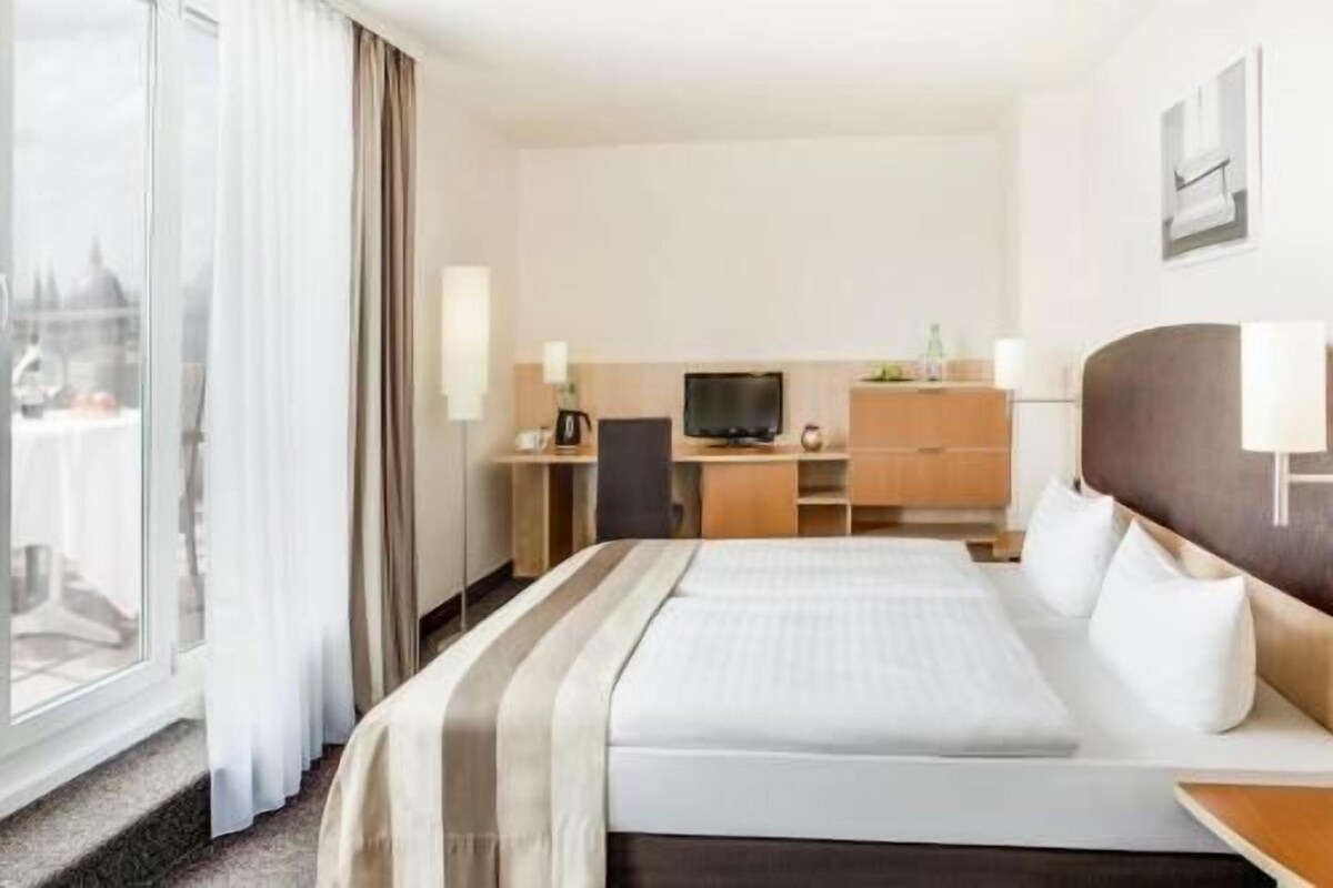 Habitación de hotel con cama, escritorio con televisor, cómoda y ventana con cortinas.