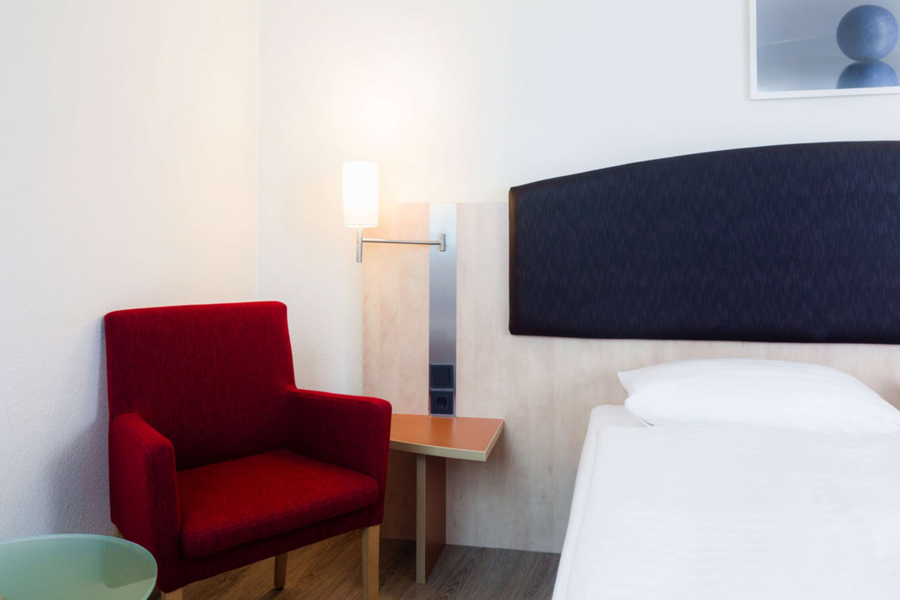 Quarto de hotel com uma cama, uma cadeira vermelha, uma mesinha, uma luminária na parede e um quadro enquadrado.