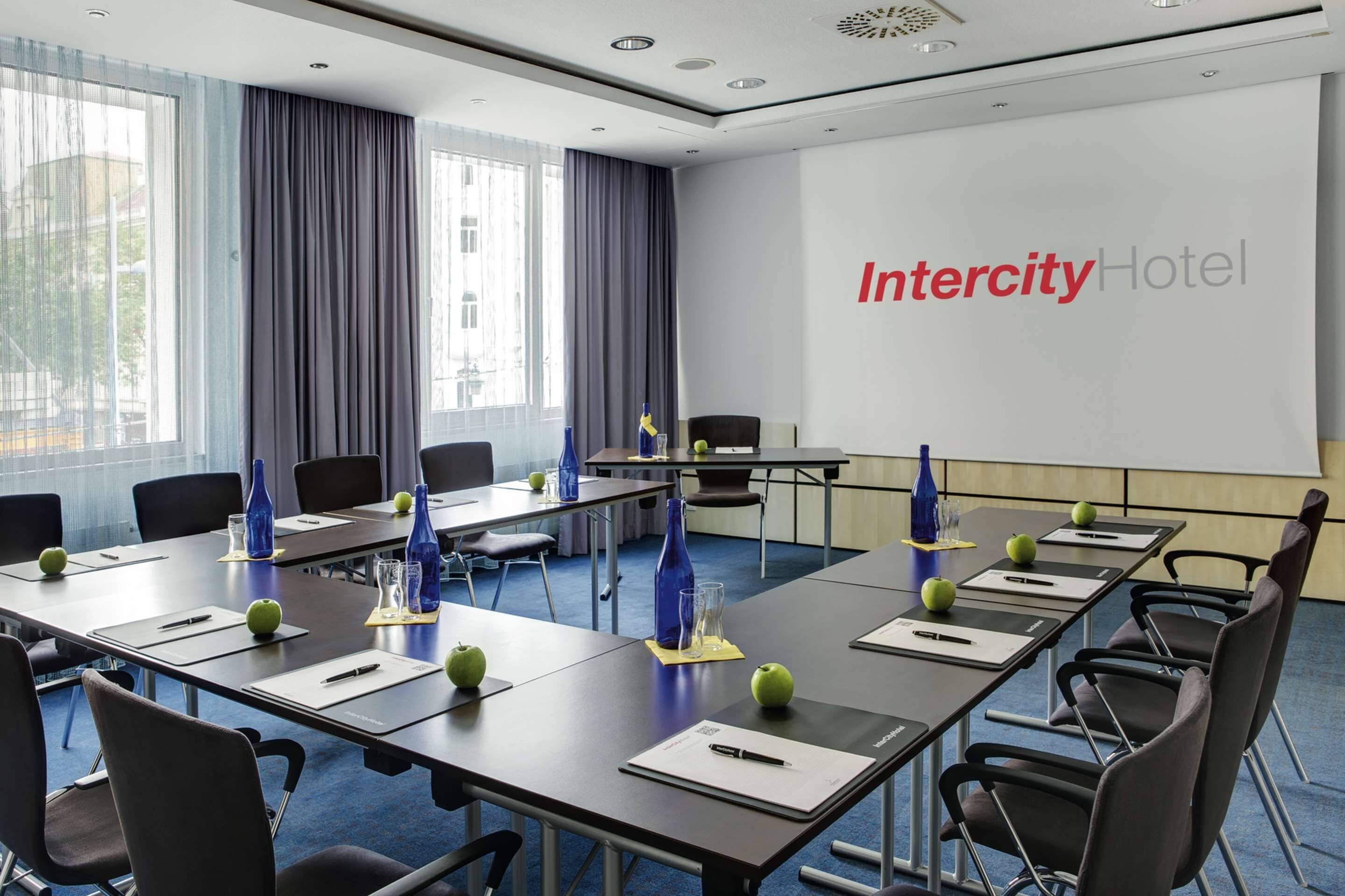 Una sala conferenze con un lungo tavolo, sedie e uno schermo proiettore che mostra "Intercity Hotel".