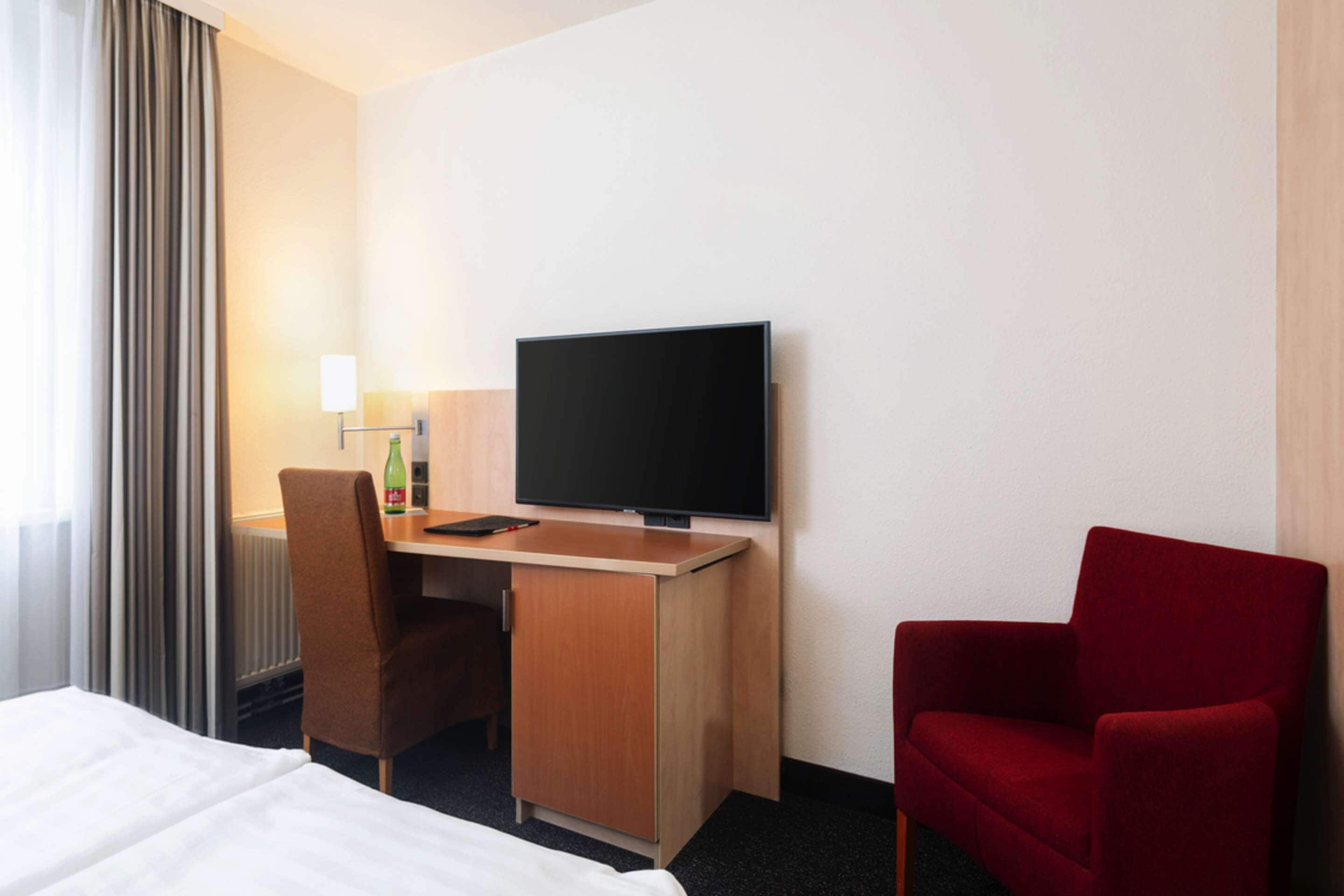 Habitación de hotel con cama, escritorio con televisor, una silla roja y ventana con cortinas.