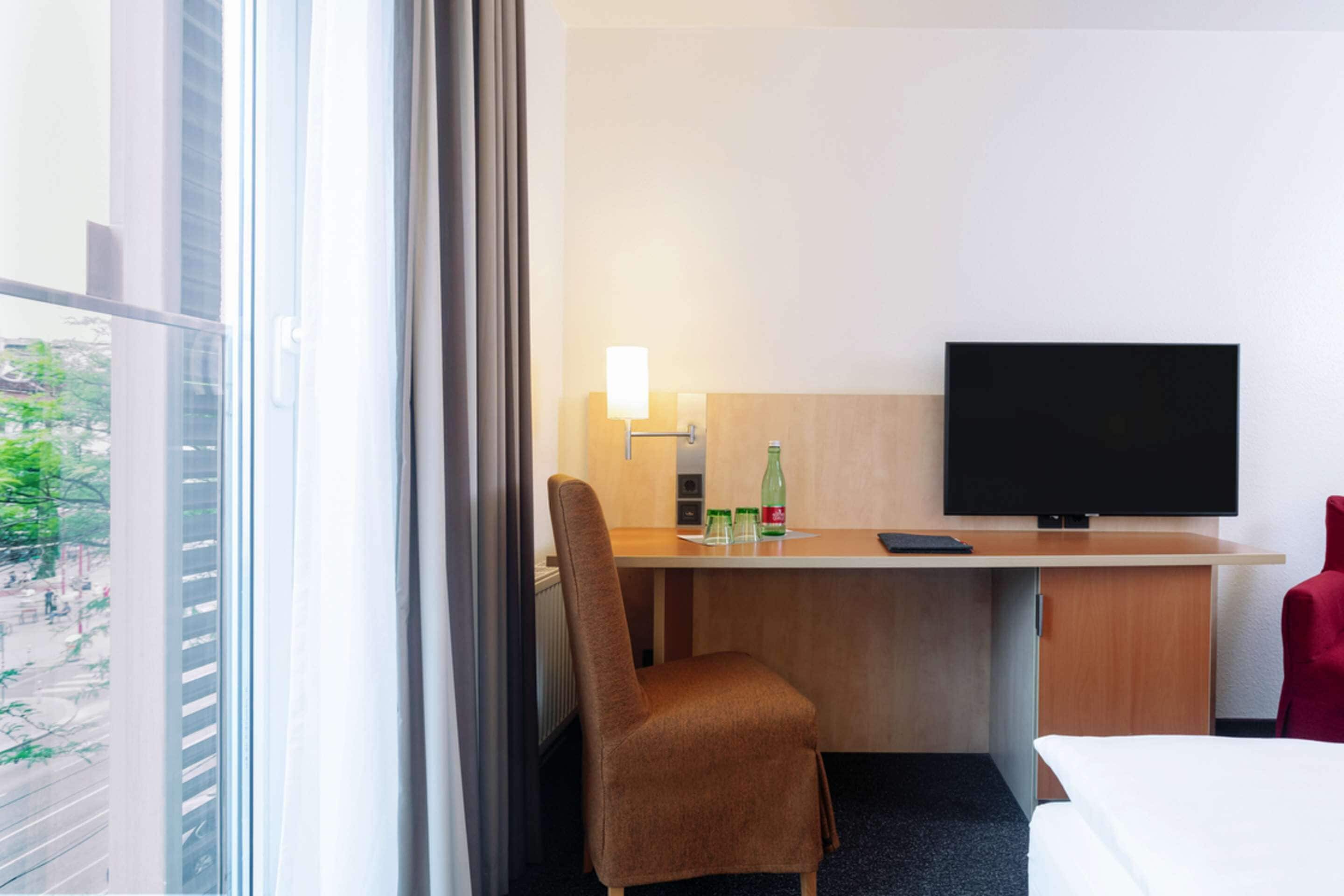 A hotel room with a bed, a desk with a TV, a chair, and a window with a view of the outside.