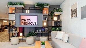 Una sala de estar moderna con una televisión grande mostrando 'ICH ON THE MOVE', un sofá gris, una mesa de centro de madera y una estantería con plantas.