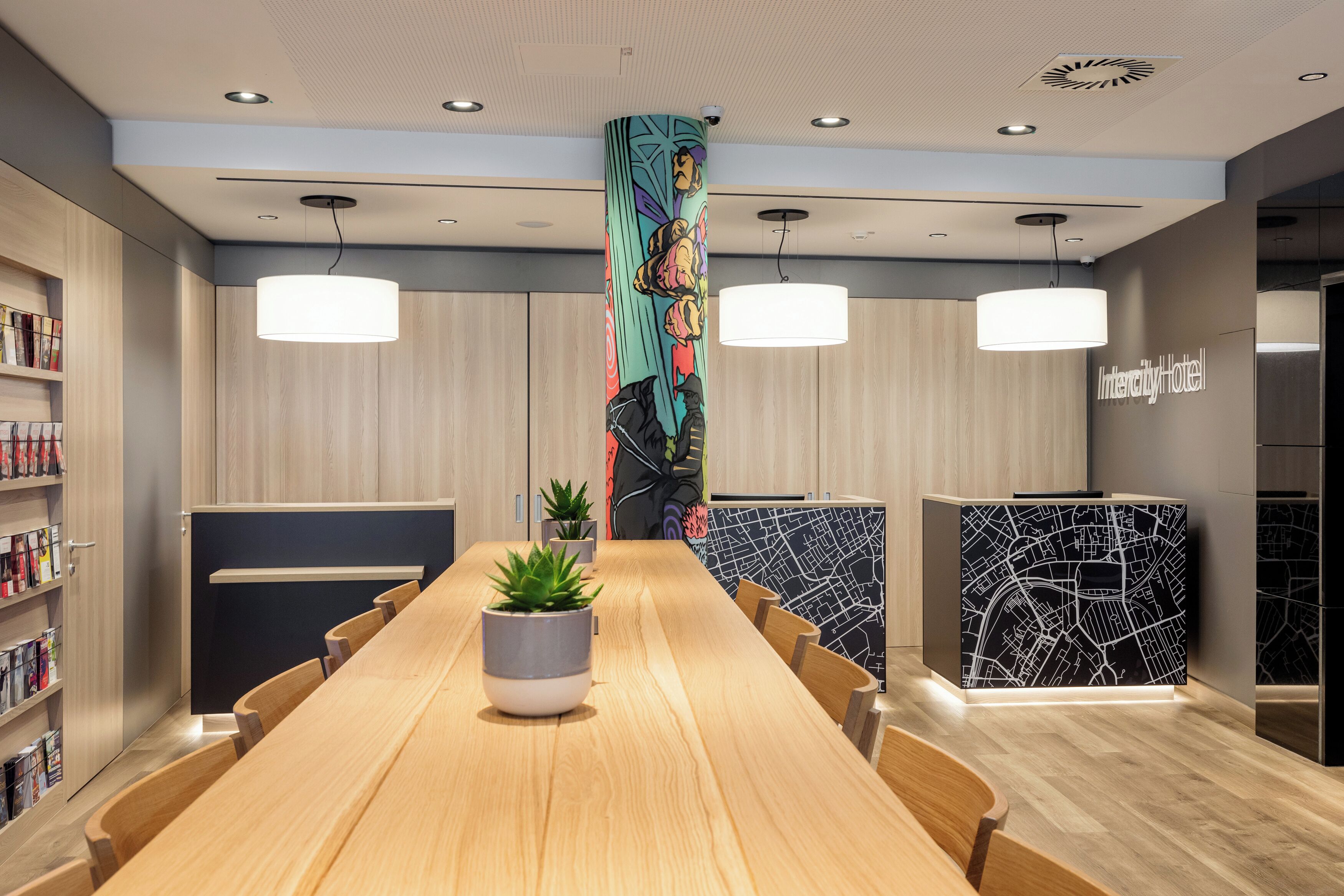Ein moderner Hotel-Empfangsbereich mit einer langen Rezeption, zwei Pendelleuchten, einer Pflanze im Topf und einer Wand mit Stadtplandesign.