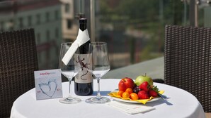 Una mesa con una botella de vino, dos copas, una bandeja de fruta y un cartel en un balcón con vistas a una ciudad que incluye un edificio con cúpula destacada.