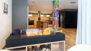 Un moderno vestíbulo de hotel con un área de estar central que incluye una mesa de vidrio con chimenea, piso de madera y un pilar de colores.