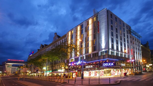 Un hotel moderno con un llamativo letrero DELKA, situado en una intersección urbana rodeado de edificios e iluminación de calle.