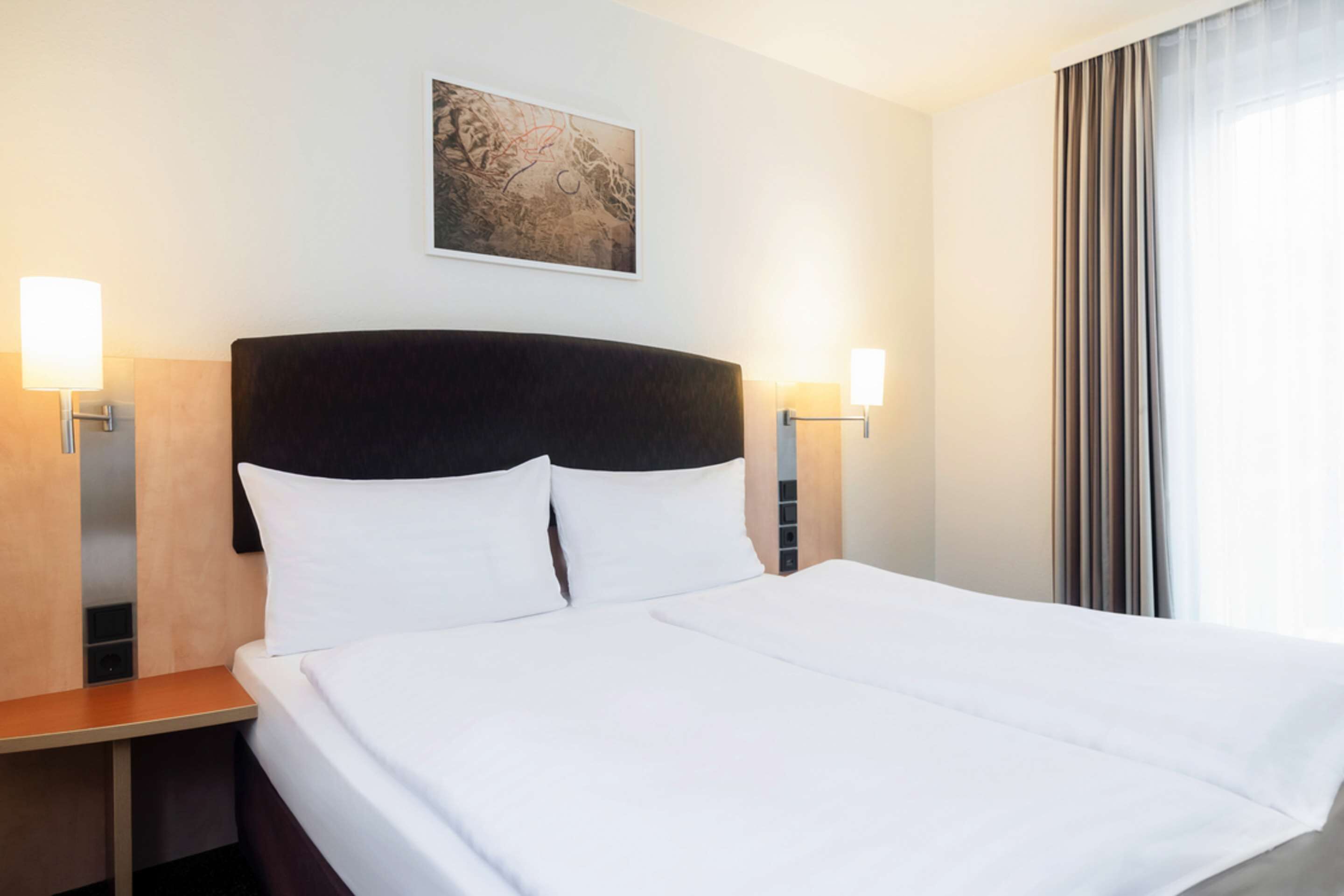 Une chambre d’hôtel avec un grand lit, deux tables de chevet, une tête de lit, une œuvre d’art accrochée au mur et une fenêtre avec des rideaux.
