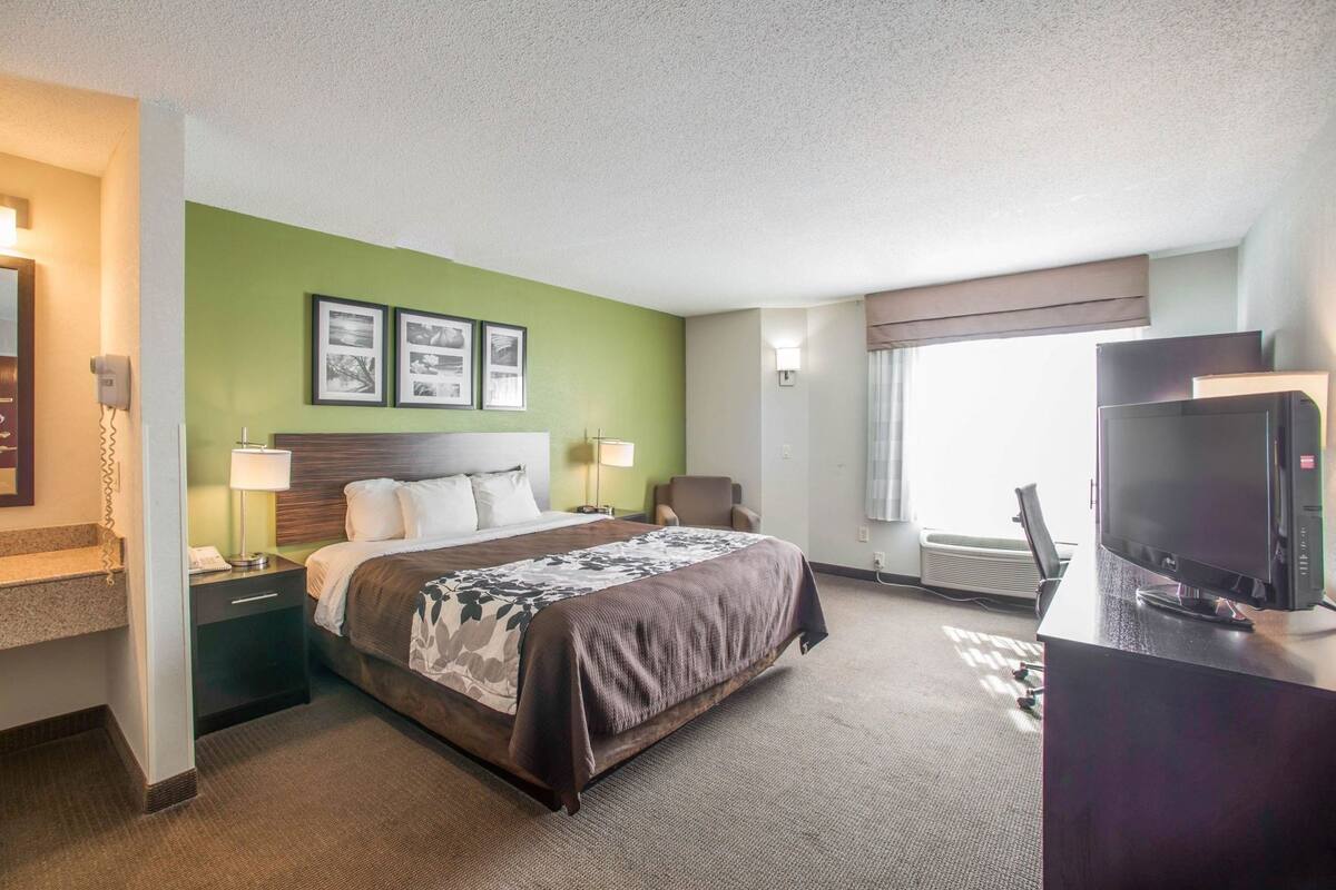 Ein Hotelzimmer mit einem Bett, Nachttischen, einem Schreibtisch mit Fernseher, einem Fenster mit Vorhängen und gerahmten Bildern an der Wand.