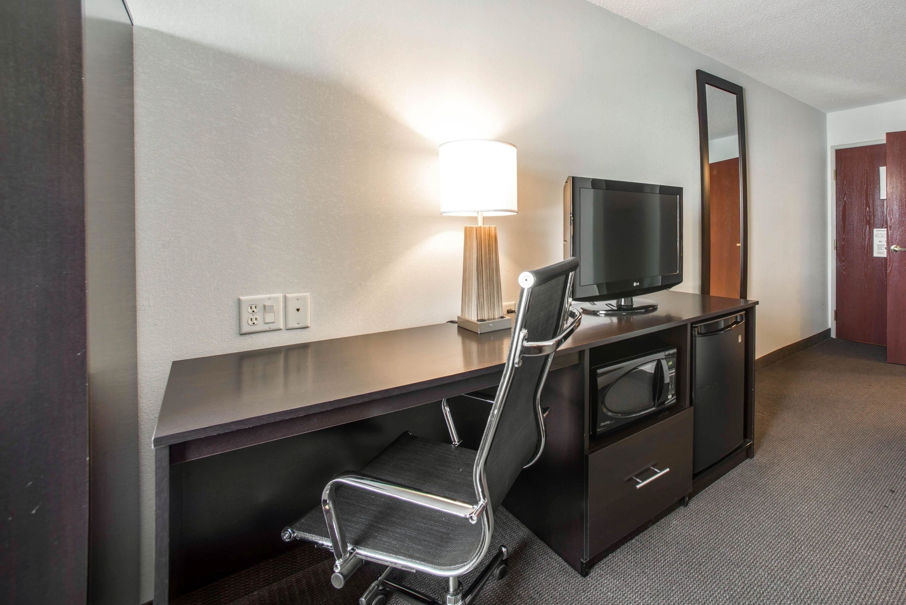 A hotel room with a desk, a chair, a television, a lamp, a mirror, and a wardrobe.