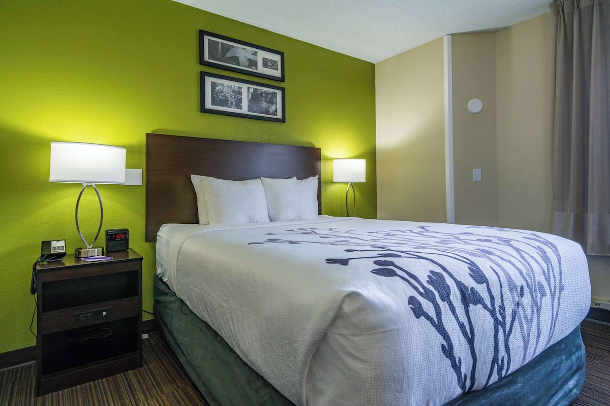 A hotel room with a bed, two bedside lamps, a nightstand with a phone and clock, and two framed pictures on the wall.