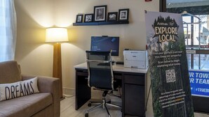 Habitación de hotel con escritorio, computadora, lámpara y sofá con almohada. Se observan cuadros en la pared y un cartel que promociona la exploración local.