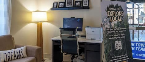 Habitación de hotel con escritorio, computadora, lámpara y sofá con almohada. Se observan cuadros en la pared y un cartel que promociona la exploración local.